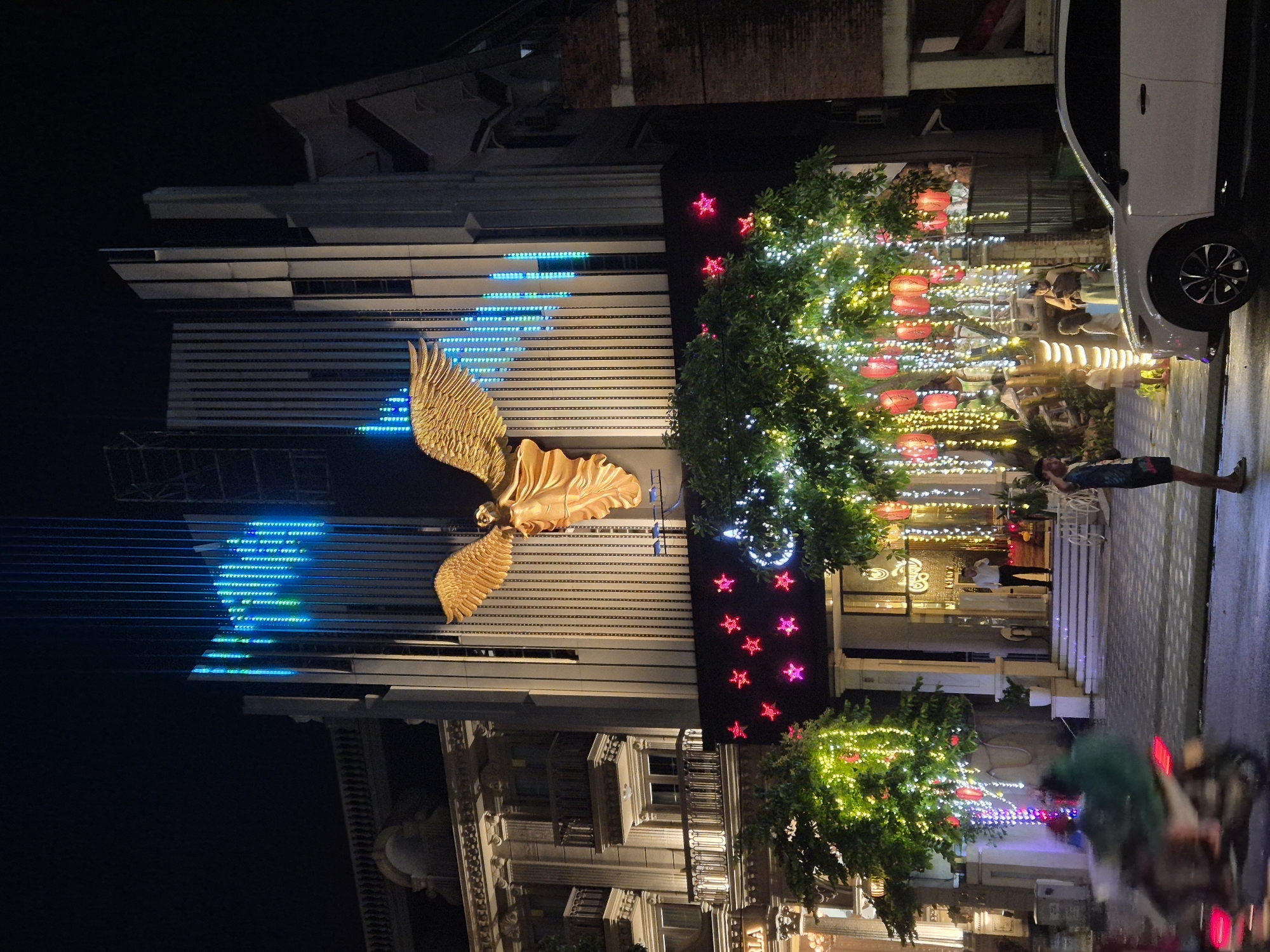 This image shows a nighttime street scene in Da Nang, Vietnam, featuring a building with a prominent golden angel sculpture. The building has a modern architectural style with vertical lines, and is decorated with blue LED lights arranged in vertical columns. Below the angel sculpture, there are red star-shaped lights. Trees in front of the building are adorned with numerous white and yellow string lights, and red lanterns are suspended from the branches. A white SUV is parked on the right side of the street. In the foreground, a person is standing and a motorcycle is blurred due to motion. The street appears to be wet, possibly from recent rain, and reflects the lights. The overall impression is of a vibrantly lit urban environment at night.
