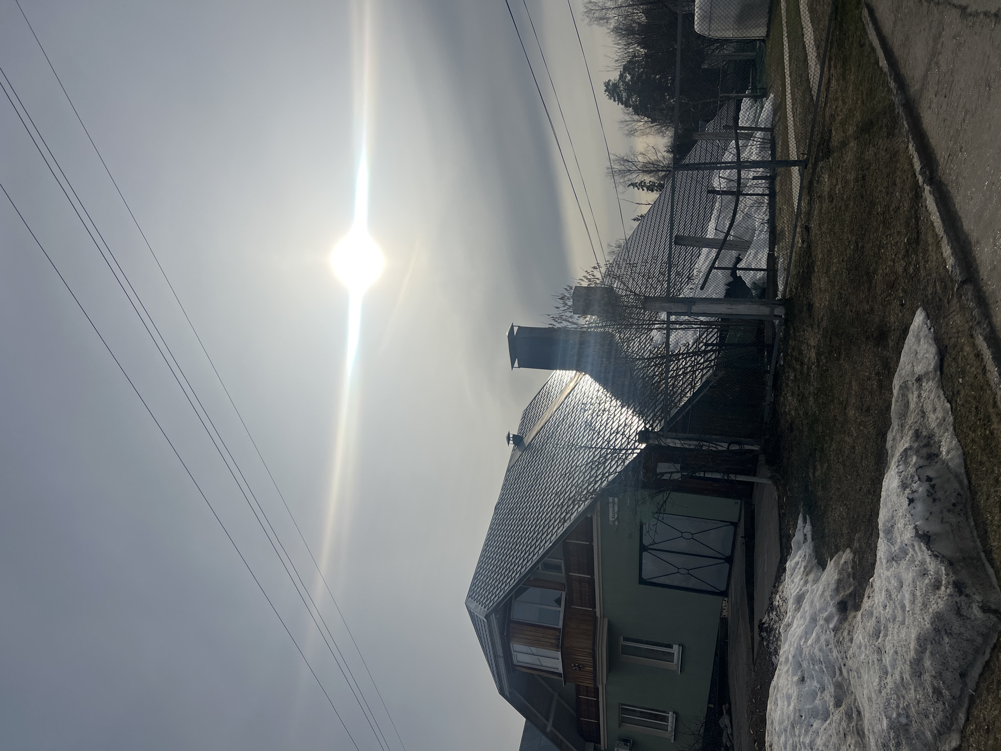 The image depicts a residential scene, potentially in a suburban or rural area. A two-story house dominates the view, featuring a dark gray tiled roof, a chimney, and light green exterior walls with dark trim. Windows are visible on both floors. The sun is high in the sky, causing a significant glare and making details difficult to discern. Overhead power lines are visible in the upper left corner of the frame. Patches of melting snow are on the ground alongside the house, which indicates it might be early spring. There is a fence behind the house and trees in the background. The location is Noviy, Russia. There is no explicit confirmation in the image that this is Noviy, Russia. No people are visible in the scene.