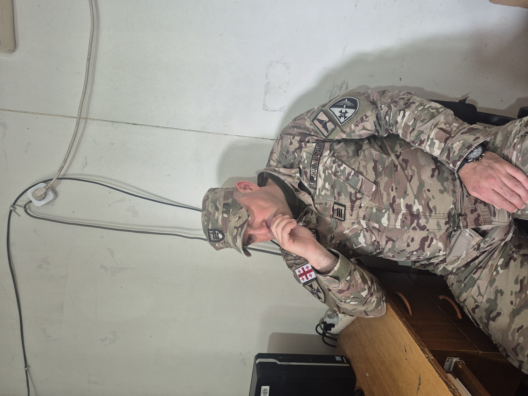 A man in military camouflage clothing is sitting on a wooden desk with his elbow resting on it, his hand near his mouth. The clothing has a Georgian flag patch on the left sleeve. A computer tower is on the floor near the desk. The background is a wall with electrical cords and an outlet. There are no identifiable details indicating the location is in Tbilisi, Georgia.