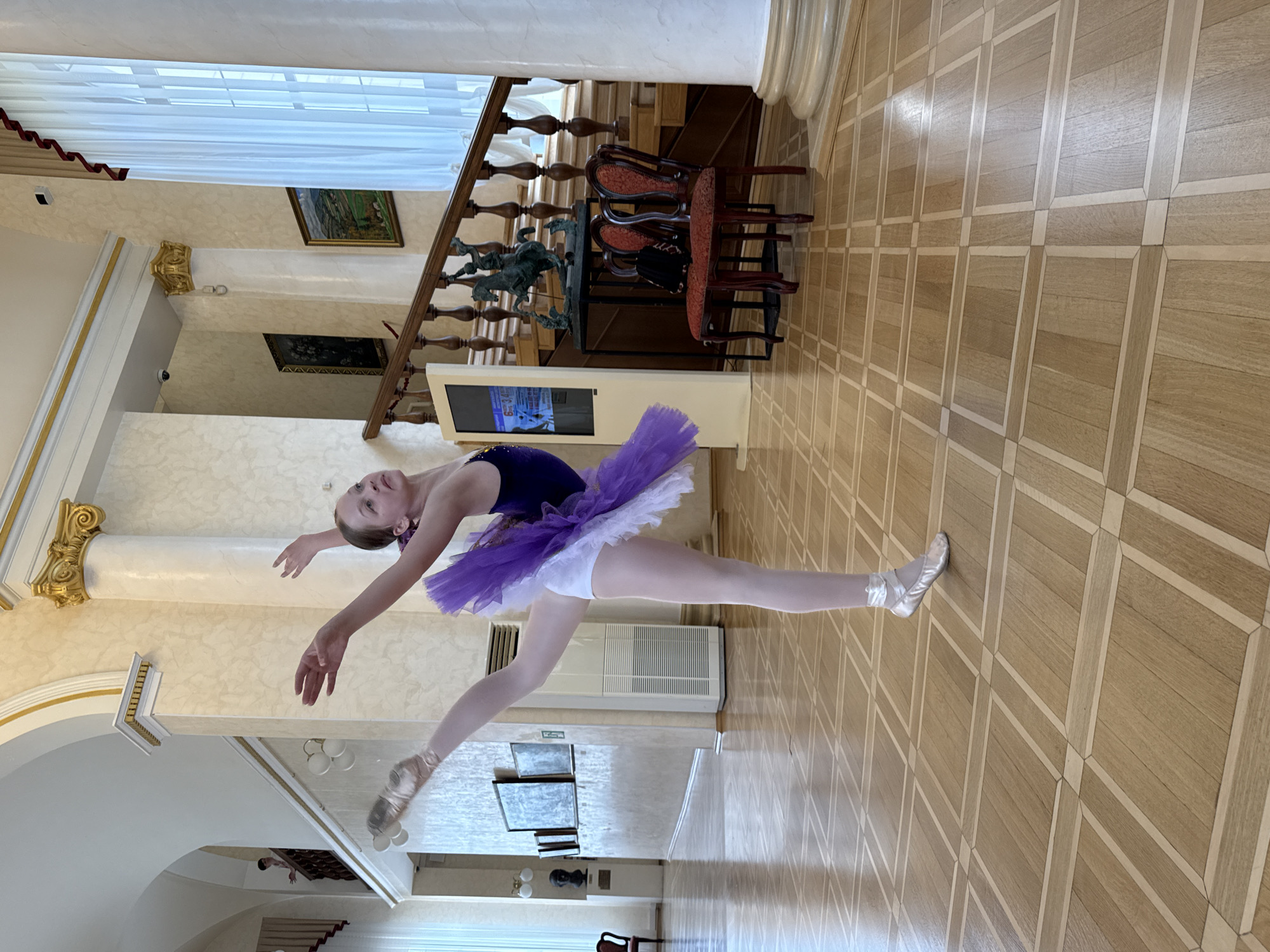 In the foyer of the Winter Theater in Sochi, Russia, a young female ballet dancer is captured in mid-pose, her left leg extended gracefully upwards and her arms outstretched. She is dressed in a dark leotard, a white tutu, and pink ballet slippers. The dancer is positioned on a polished wooden floor with a geometric inlay pattern. In the background, a grand staircase with ornate wooden railings is visible, leading to an upper level. Paintings adorn the walls, and large white curtains hang near a window. Two antique red chairs with dark seats sit to the right of the staircase. A digital display screen stands to the left of the staircase. The architecture features tall, fluted columns and ornate gold detailing on the archways and capitals. Sunlight streams through the curtains, illuminating the scene.