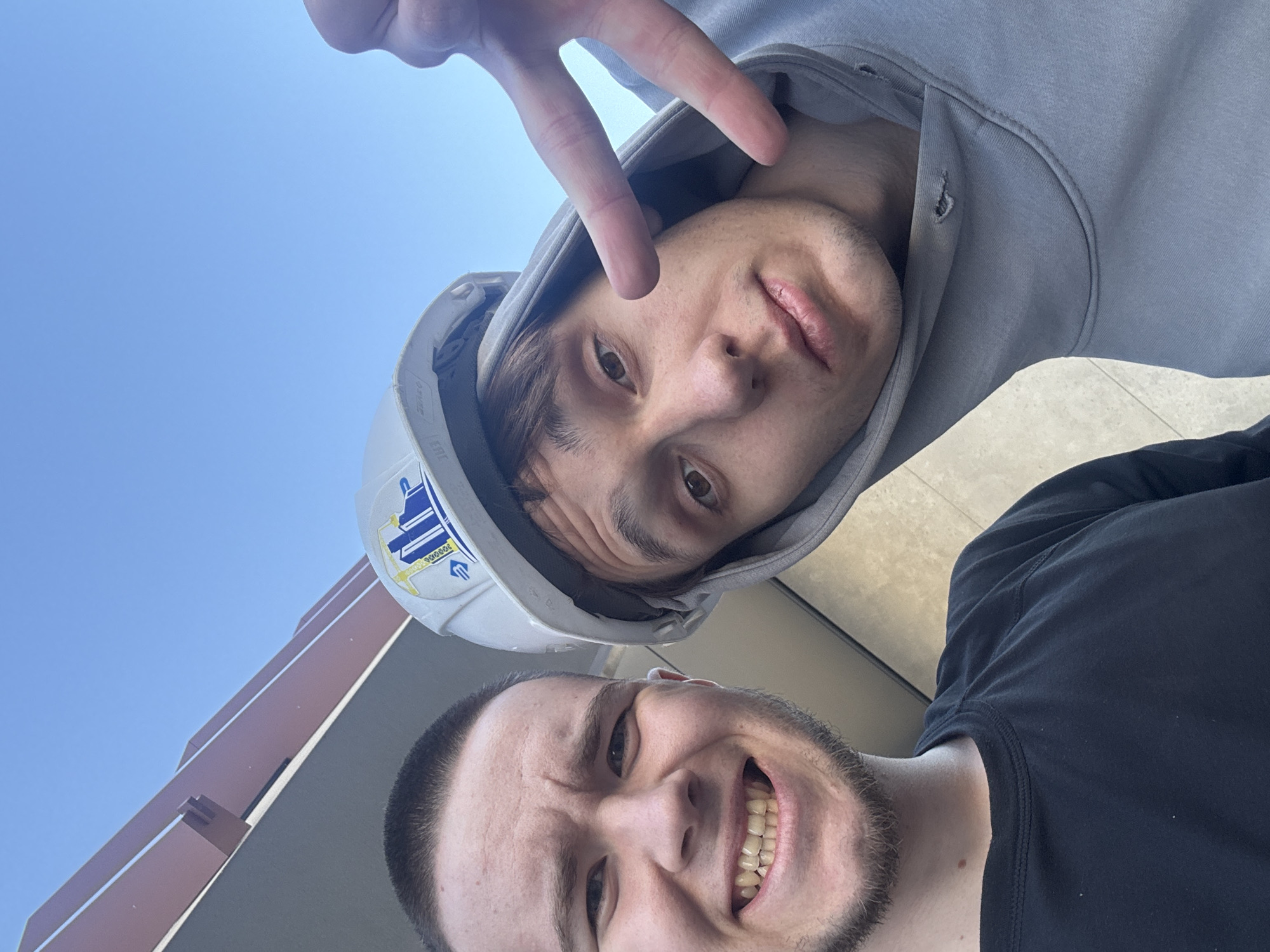 Two Caucasian males are visible in a selfie. The top male is wearing a gray hooded sweatshirt and a white hard hat with a blue and yellow emblem, making a "peace" sign with his fingers. The bottom male is wearing a black shirt and smiling. The sky is light blue. The lower-left portion of the image shows a brown building. The location is likely Kazan, Russia based on the emblem on the hard hat.