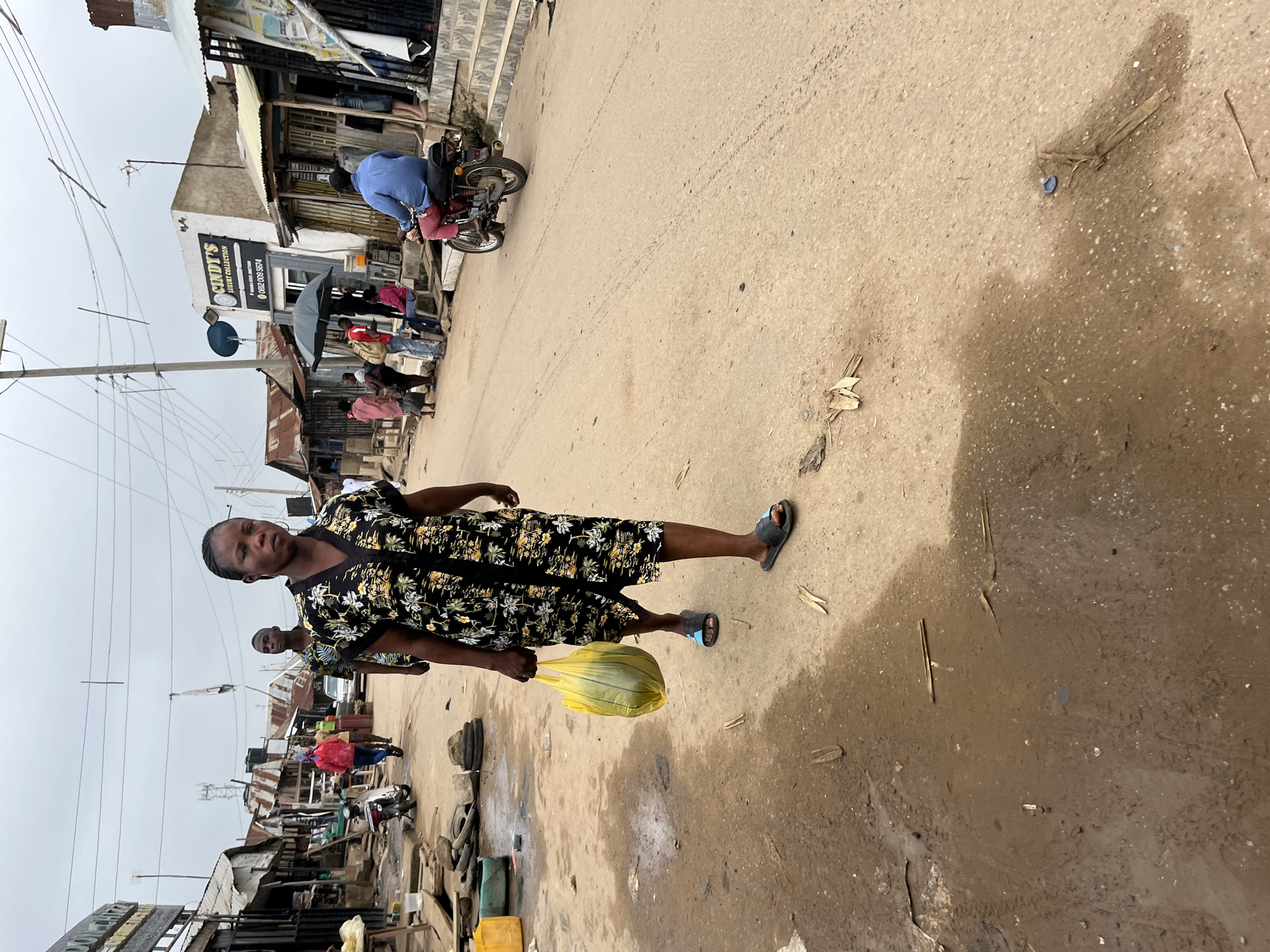 The image depicts a street scene in Abuja, Nigeria, characterized by an unpaved, dirt road with puddles of water. Several structures, likely shops or homes, line the street, constructed from corrugated metal and rough materials, with some appearing weathered. Utility wires are visible overhead, strung between poles.
In the foreground, a woman walks towards the camera, holding a yellow plastic bag in her left hand. She is wearing a dark, floral-patterned dress and dark flip-flops. Her gaze is directed forward. Behind her, another person, a man, is partially visible, also walking away from the camera. Further down the street, several other individuals are present. One person is riding a motorcycle, wearing a blue shirt and pink pants. Others are standing or sitting near the roadside, some gathered under an umbrella.
Objects scattered on the road include discarded tires, wooden debris, and a blue bottle cap. The overall impression is one of a busy, informal market or residential area, with signs of daily life and economic activity. The sky is overcast, suggesting a cloudy or potentially rainy day.