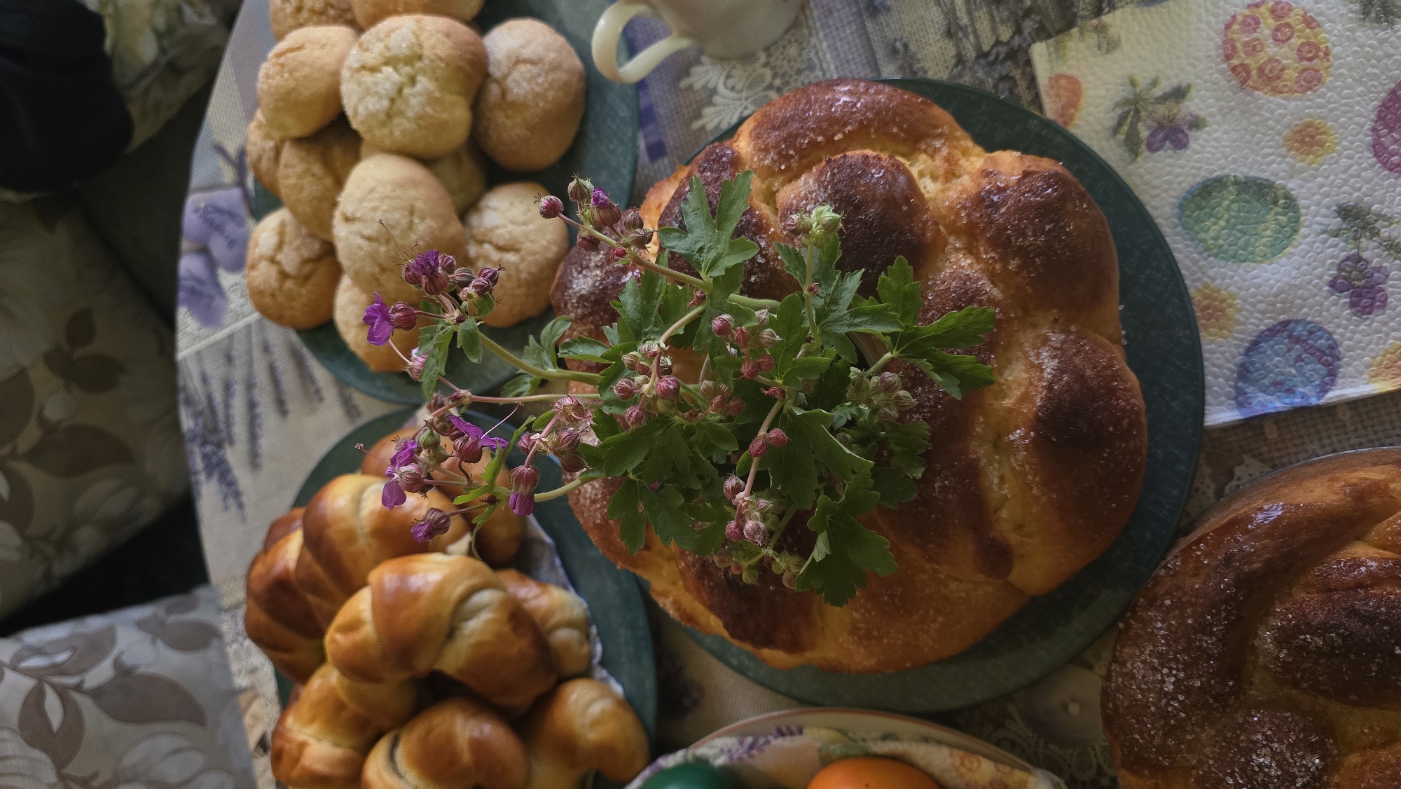 The image shows a table spread with baked goods, likely in a home setting in Stara Zagora, Bulgaria, given the specific types of bread present. There are two large braided loaves, characteristic of traditional Bulgarian Easter bread (kozunak), decorated with a sprig of wild geranium. Adjacent to these are plates filled with small round rolls and crescent-shaped pastries. Beneath the kozunak is a partial view of a basket containing colored Easter eggs (pisanitsa). A napkin with Easter egg designs adds to the festive theme. The composition suggests a celebratory meal or gathering, common during Easter in Bulgaria.