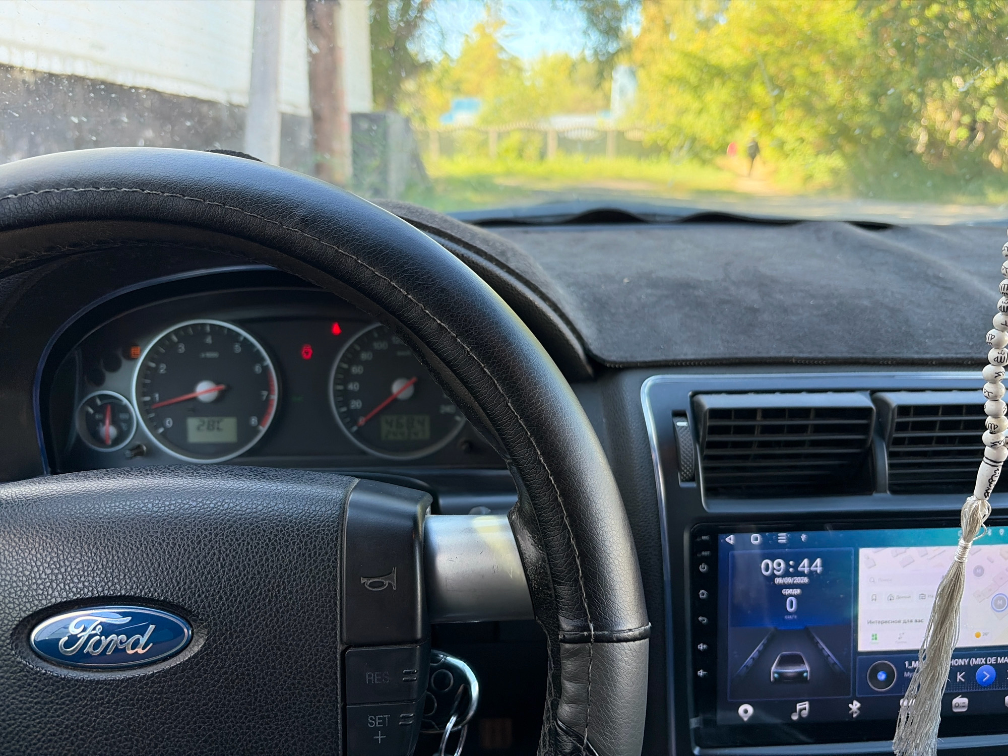 The image provides an interior view of a car, likely a Ford, as indicated by the logo on the steering wheel. The dashboard features a steering wheel with control buttons and the Ford emblem. To the right of the steering wheel, a car's infotainment system is visible, displaying the time as 09:44, the date 09/09/2026, and the speed as 0 km/h. The infotainment screen also shows a navigation interface with a map and an indication of "Interesting for you." A string of prayer beads hangs from the rearview mirror area. The car's instrument cluster, including a speedometer and tachometer, is partially visible behind the steering wheel. The windshield shows an outdoor scene with trees and a fence in the background, suggesting a rural or suburban setting. There is a hint of a building on the left. The overall impression is of a parked vehicle. The location is identified as Железинка ауылдық округі, Kazakhstan.