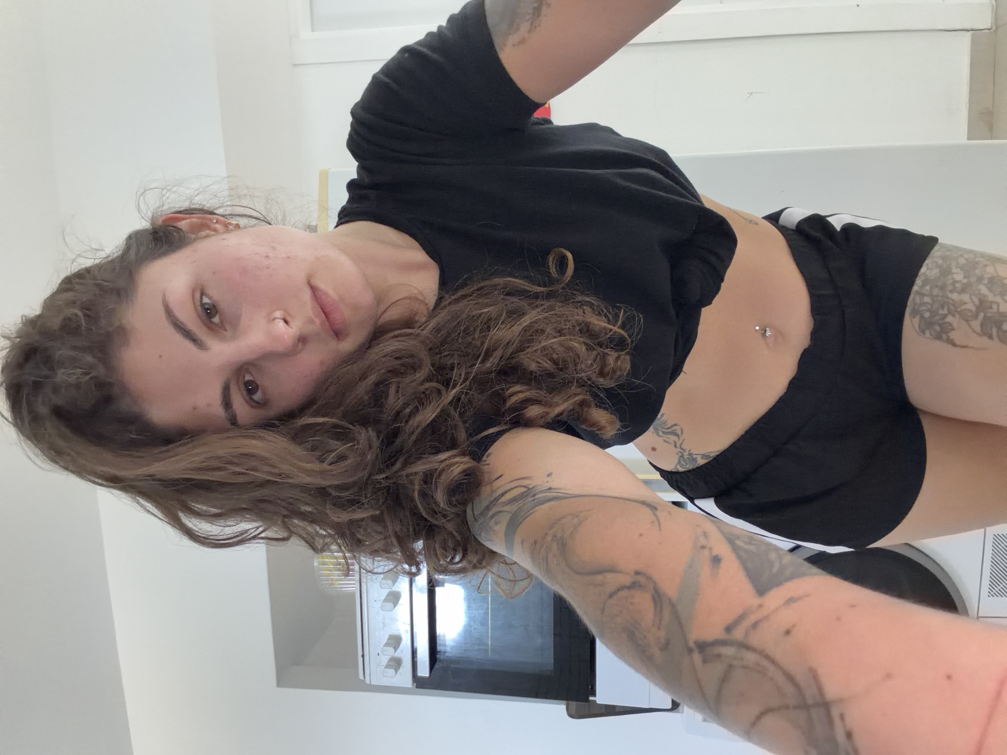 A woman is indoors, possibly in a kitchen, wearing a black t-shirt, black shorts, and has body piercings. The woman also has a tattoo on her chest, and a tattoo on her arm. There is an oven in the background, and the setting may be a kitchen. It is unknown if the location is in Kiryat Bialik, Israel.