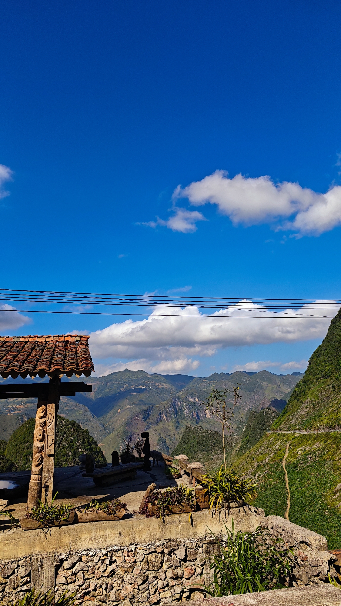 The photograph depicts a vista from the Đồng Văn Commune, Vietnam. The foreground features a stone-walled patio with planters. On the patio is a wooden structure with a tiled roof, adorned with carved details. Beyond the patio is a mountainous landscape with roads and foliage. The sky is blue with scattered white clouds. Overhead, power lines extend across the frame.