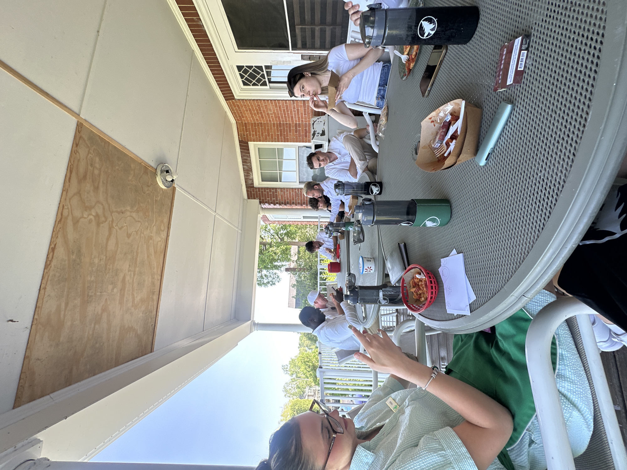 A group of eight people are gathered around a round table on an outdoor porch, eating and socializing. The porch is covered by a wooden ceiling with exposed beams. The individuals are dressed casually, and most are looking at their phones or food. Food containers, bottles, and napkins are scattered on the table. The background shows trees and a brick building. The scene suggests a casual outdoor dining experience.