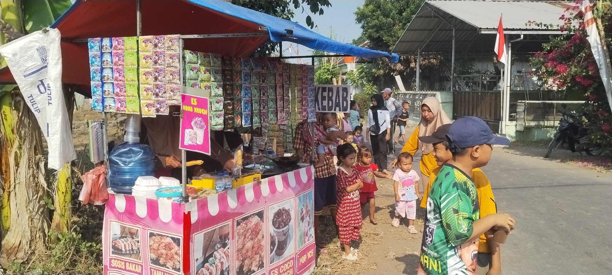 This image captures a street-side food stall in Yosowilangun Kidul, Indonesia, on a sunny day. The stall, adorned with a blue tarp and a pink and white striped awning, displays a variety of packaged snacks and food items. Prominently featured are images of "Sosis Bakar" (grilled sausage) and "Es Coco Kruch" (a type of ice dessert), suggesting these are popular offerings. A large blue water dispenser is visible, along with stacks of white disposable containers. Several people are present, including a woman in a hijab speaking with children, a child being held by another person, and a group of children standing near the stall. The background shows a residential area with houses and trees. An Indonesian flag flies from a pole attached to a nearby building. A motorcycle is parked on the side of the road. The overall scene suggests a typical day at a local marketplace or roadside vendor.