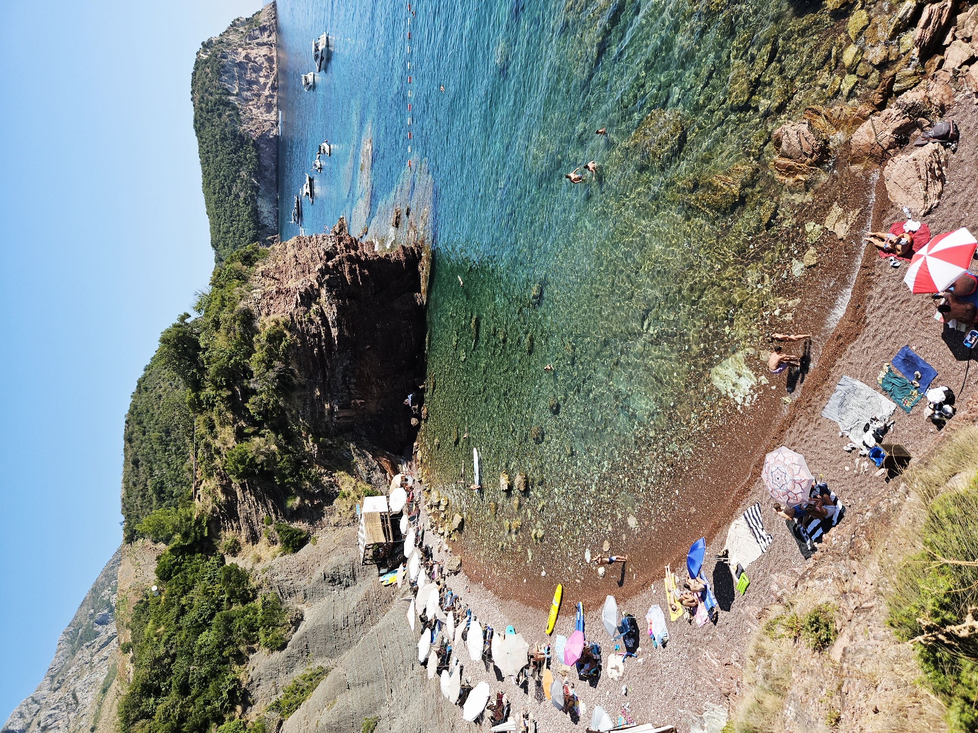 This is an aerial, high-angle, eye-level shot of a rocky, pebbled beach and clear, turquoise water in Sveti Stefan, Montenegro. The beach is lined with numerous white beach umbrellas, some of which are open, providing shade to sunbathers. Several people are visible on the beach, some lying on towels or lounging under umbrellas, while others are walking or standing. In the water, several individuals are swimming, and one person is on a stand-up paddleboard. A small wooden beach bar or shack is situated at the edge of the beach. The water is transparent, allowing the rocky seabed to be clearly seen. Boats are anchored in the deeper water, and a cliff face covered in lush green vegetation rises behind the beach. The sky is a clear, bright blue, indicating a sunny day. The scene depicts a tranquil and scenic coastal resort environment.