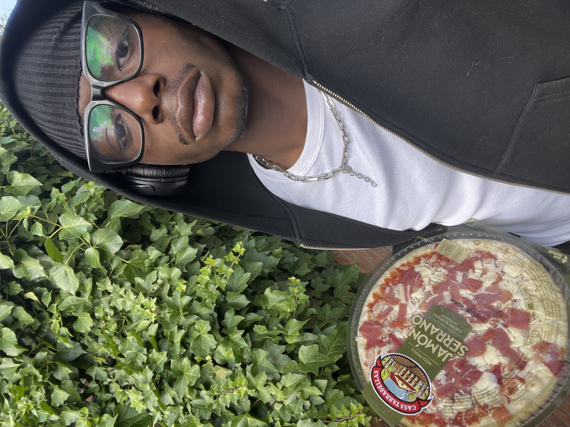 A medium shot shows a person holding a pizza in front of a dense ivy hedge. The person, a black male, wears a black beanie, black-framed glasses, and headphones. He is also wearing a white t-shirt and a black hooded jacket. A silver chain necklace is visible. The pizza is a pre-packaged "Casa Tarradellas Jamón Serrano" pizza. The ivy is lush and covers a wall or ground, suggesting a park or garden setting. The image is slightly rotated.