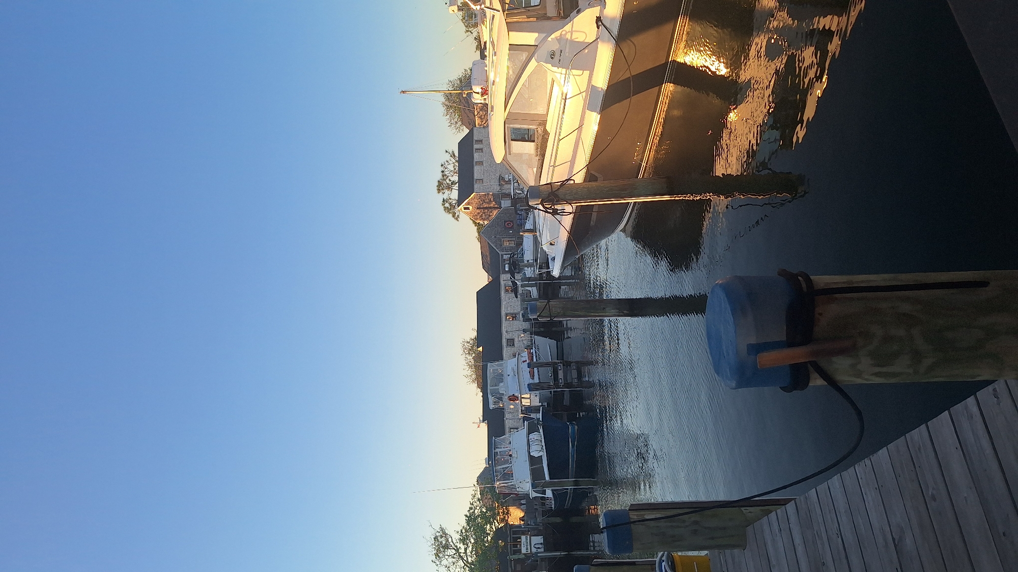 A vertical, eye-level, outdoor, full shot captures a serene harbor scene on Nantucket, USA, bathed in the warm glow of late afternoon sun. The sky is a clear, pale blue, transitioning to a slightly lighter hue at the horizon. In the foreground, a wooden dock leads the eye towards the water. Mooring posts, some capped with blue, rise from the dark water, their weathered textures contrasting with the smooth, reflective surface.
Several boats are docked in the harbor. To the left, a white boat with a blue hull is prominently featured. To its right, a white boat with a dark hull and a light-colored cabin is illuminated by the sun, its reflection shimmering on the water. Further in the background, more boats and a cluster of traditional Nantucket-style buildings with gray shingles and dark roofs are visible. The architecture suggests a historic coastal town. The overall atmosphere is peaceful and tranquil, with the setting sun casting long shadows and highlighting the textures of the boats and wooden structures. No people are visible in the frame.