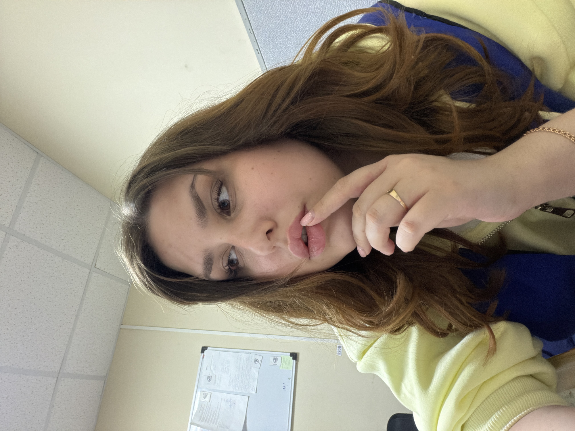 A young woman with brown hair, wearing a yellow sweater with a blue undershirt, is posing indoors in what appears to be an office or institutional setting. She is wearing a gold ring and a gold bracelet. A whiteboard with papers attached is visible on the wall. The ceiling is a standard grid pattern. The picture was taken in Chaikovsky, Russia.