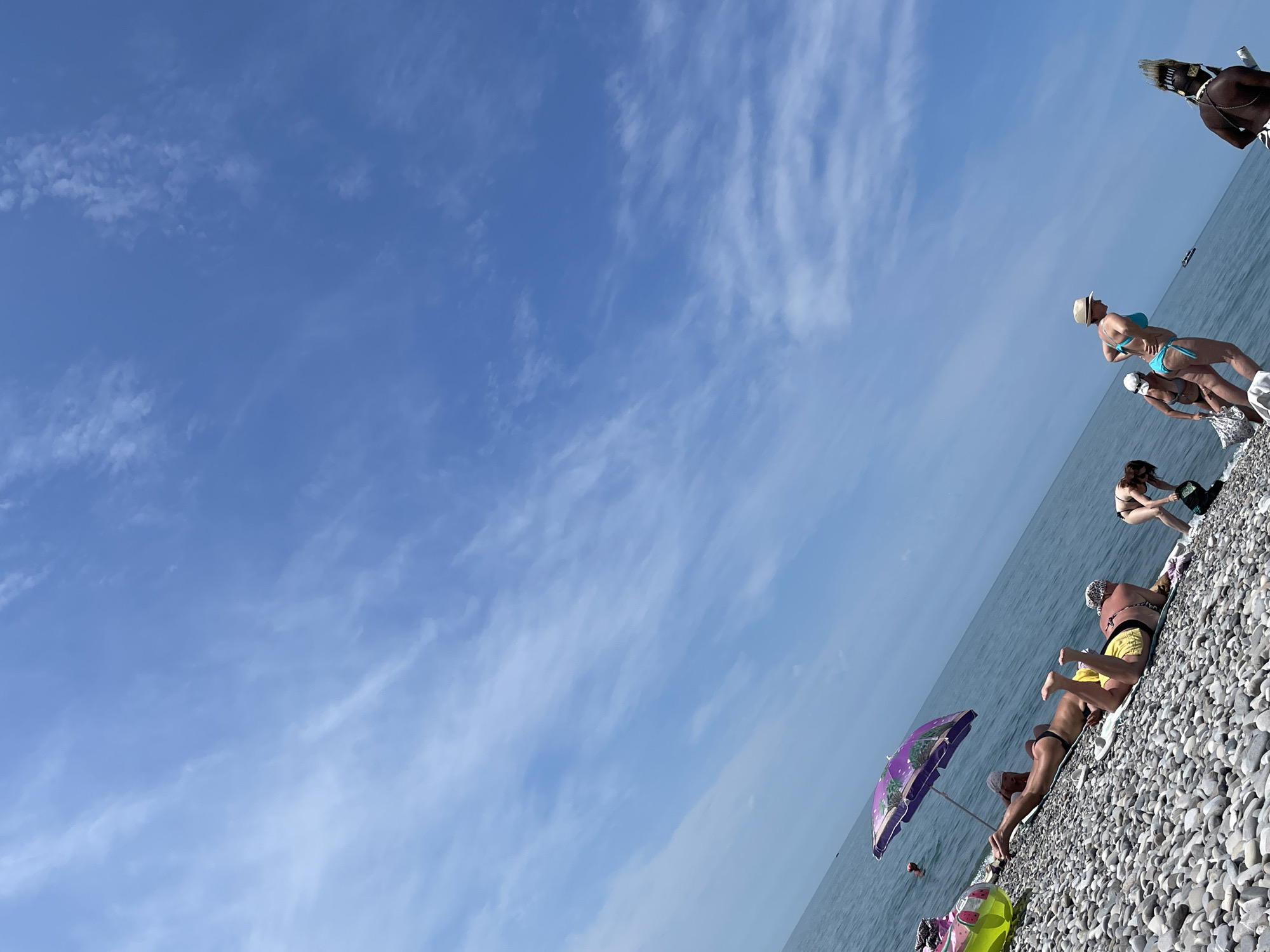The image depicts a sunny day at a pebble beach. The sea stretches out to the horizon under a blue sky with scattered white clouds. Several people are present on the beach. In the foreground, a person in a black bikini and another in yellow swim trunks are sunbathing on mats. A purple umbrella with a tropical print is set up nearby. To the right, a woman in a blue bikini and a straw hat is walking, while further to the right, a person wearing a headdress that resembles a tribal costume stands on the shore. A few other individuals are visible in the background, some in the water and others on the beach. The overall scene suggests a relaxed atmosphere at a beach resort.