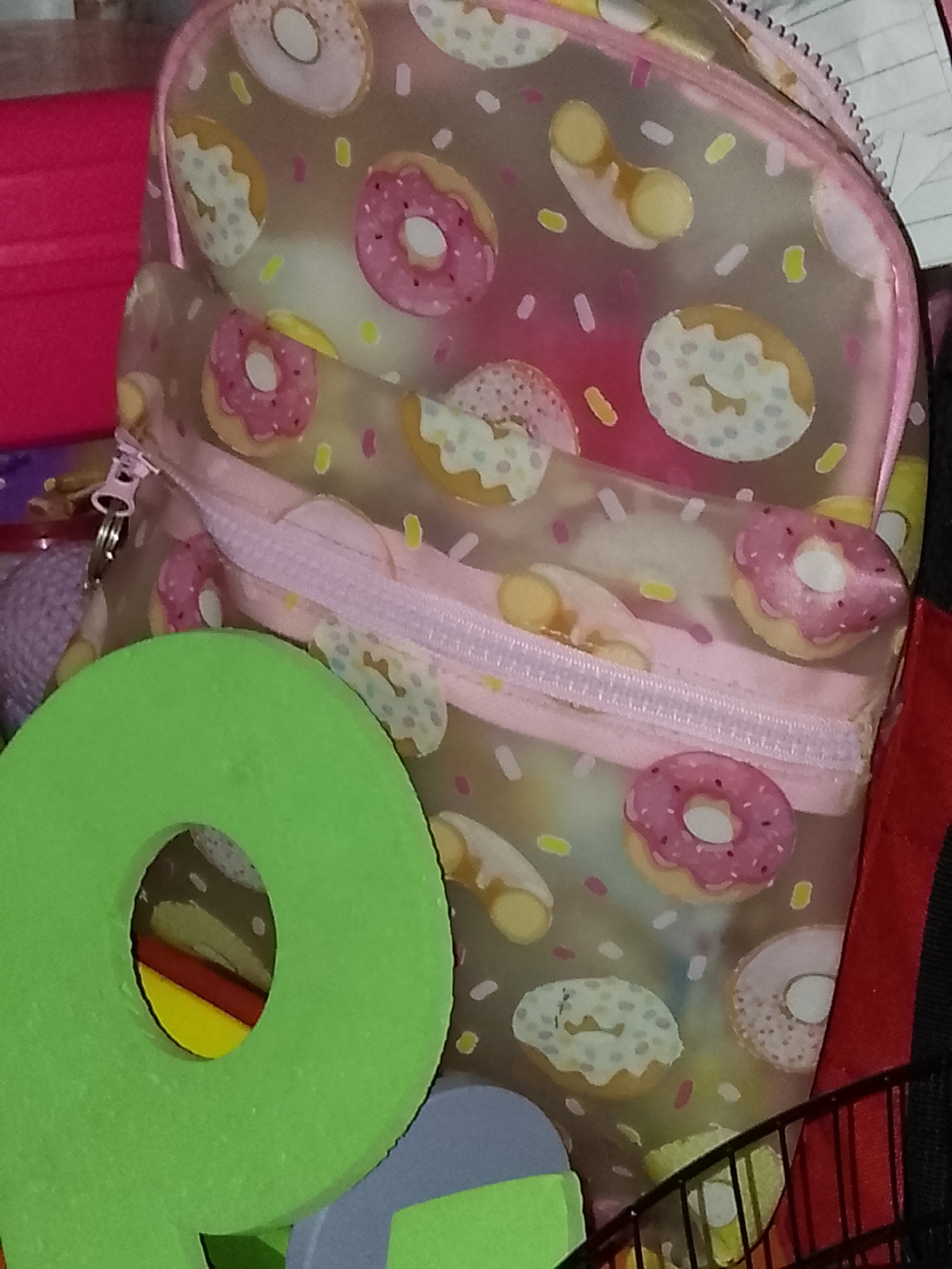 The image presents a close-up, static view of various objects in an indoor setting. The central subject is a clear or translucent small bag, possibly a pencil case, adorned with a playful pattern of donuts. The donuts are illustrated with pink, white, and yellow frosting, complete with colorful sprinkles. A pink zipper with a metallic pull is prominently visible on the bag's front pocket.
In the foreground, partially obscuring the bag, is a large, bright green foam object, which appears to be a letter 'Q' or a similar shape with a circular cutout. Through this cutout, hints of yellow and red items can be seen in the background. To the left of the donut-patterned bag, a textured purple object, possibly a ball of yarn or a soft toy, is partially visible. Further back and to the right, a red object, resembling a container or fabric, is present, along with a black, grid-like structure that might be a shelf or railing.
No specific activity or event is taking place; the image captures an arrangement of items. The setting appears to be an organized storage area or a child's space. There is no visible text within the image.