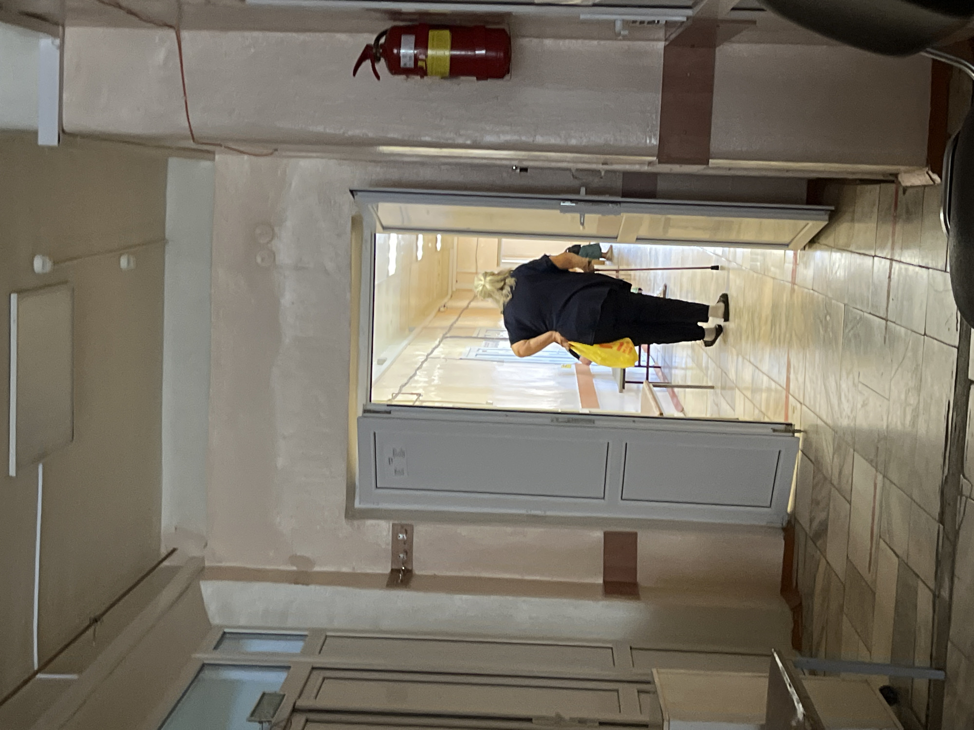 The image depicts an indoor hallway, likely in a medical facility, with a woman using a cane walking away from the camera and towards an open doorway. The hallway has tiled floors and beige walls, with a red fire extinguisher mounted on the right wall. Fluorescent lighting is visible on the ceiling. The woman is carrying a yellow bag and is dressed in dark clothing. The scene appears to be an ordinary day in a public space.