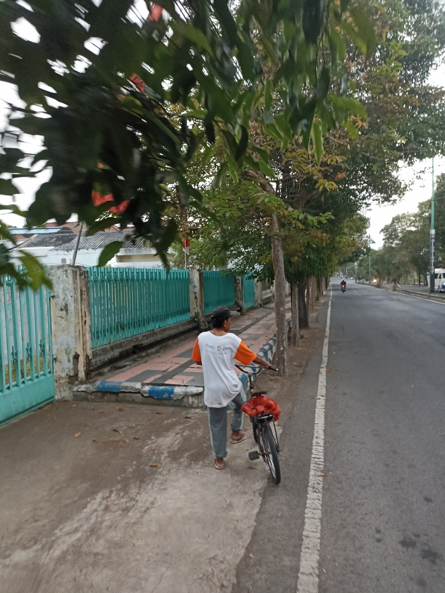 A person walks with a bicycle on a sidewalk next to a road. The person is wearing a white t-shirt with an orange collar and grey pants, and a black hat. The bicycle has a red bag on the back. The sidewalk has a patterned surface with blue and white accents. A teal colored fence runs along the sidewalk. Trees line the sidewalk and the road. The road appears to be a main street with some traffic. The location is Mojopanggung, Indonesia.