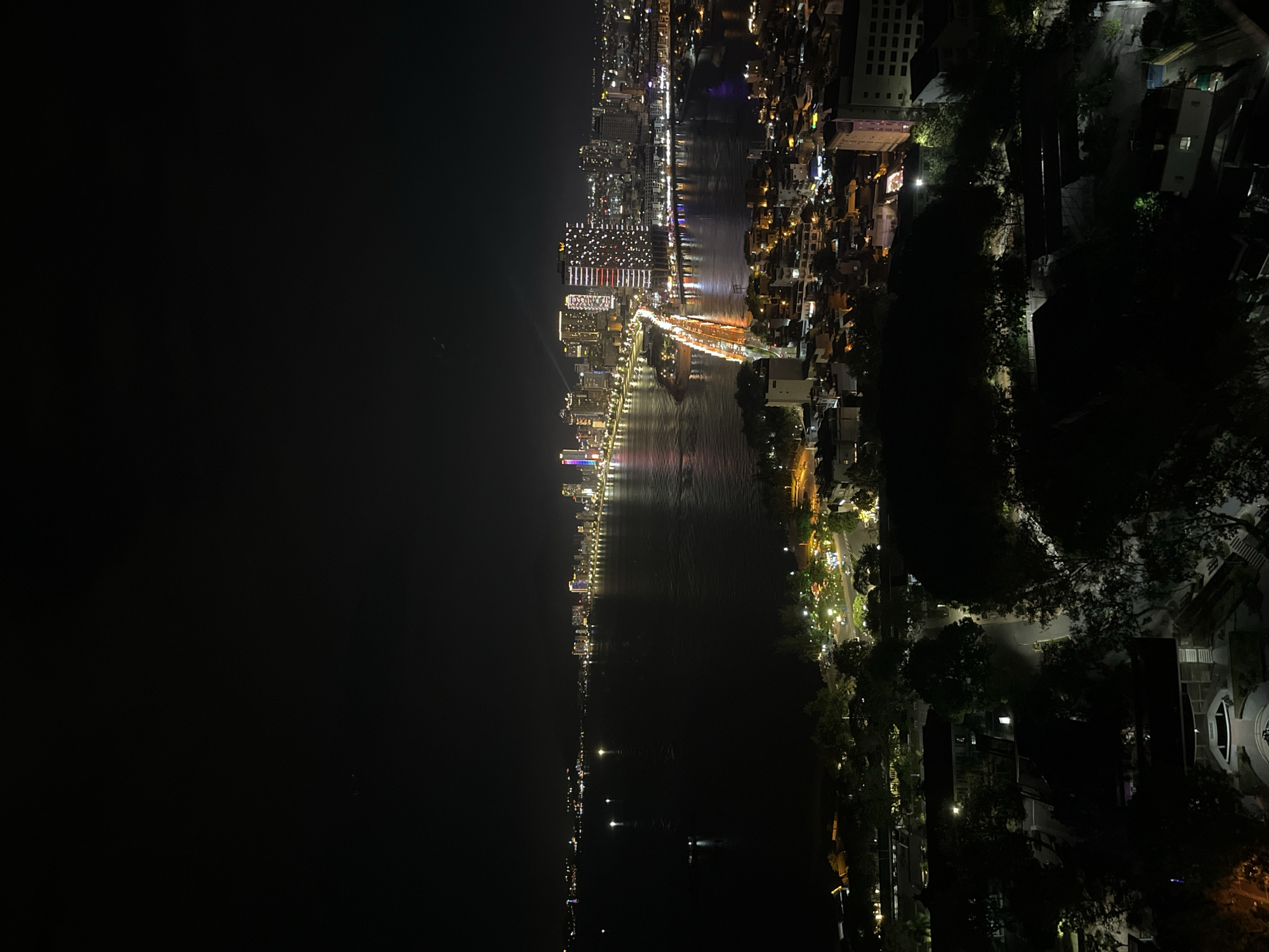 A high-angle, nighttime photograph shows a city at night. The image is oriented vertically. A river runs down the center, flanked by urban development on either side. The left side features a dense collection of buildings illuminated by bright lights, suggesting a city center or commercial district. Lights reflect off the water, creating shimmering lines. On the right, the land is covered with greenery, which is interspersed with buildings and roads lined with streetlights. The sky is dark and clear.