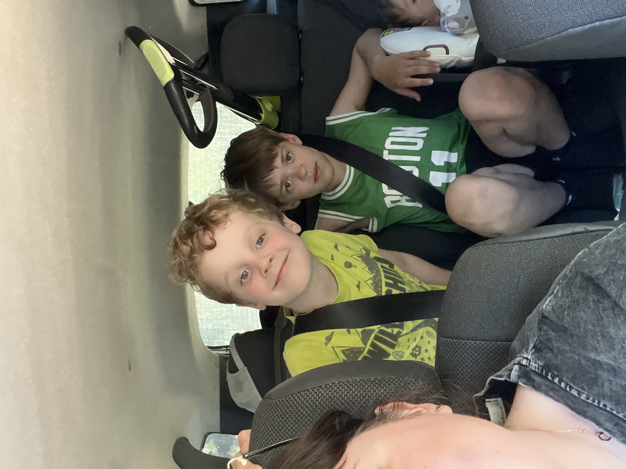 The image depicts three children in the back of a vehicle. The child closest to the camera has light skin, curly red hair, and is wearing a neon green shirt. They are smiling. An older child is behind them, wearing a green Boston Celtics jersey. They are looking at the camera with a neutral expression. A third child, possibly an infant, is further in the background lying on a pillow. All three children appear to be wearing seatbelts. An adult is partially visible in the foreground with dark hair and glasses. The interior of the vehicle is visible. The image does not depict anything to suggest it was taken in Kirillovka, Russia.