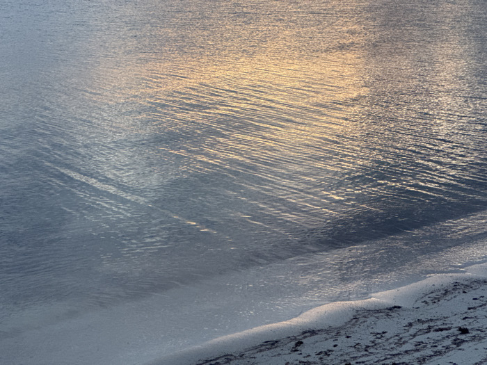 A close-up showcases the edge of a body of water meeting the shore. Gentle, rhythmic ripples and small waves texture the water's surface, reflecting a soft, golden light. Near the shoreline, the water becomes clearer, revealing a sandy beach that transitions from wet sand to drier, darker sediment. The overall mood is tranquil and serene, highlighting the subtle textures and interplay of light on the water and sand.