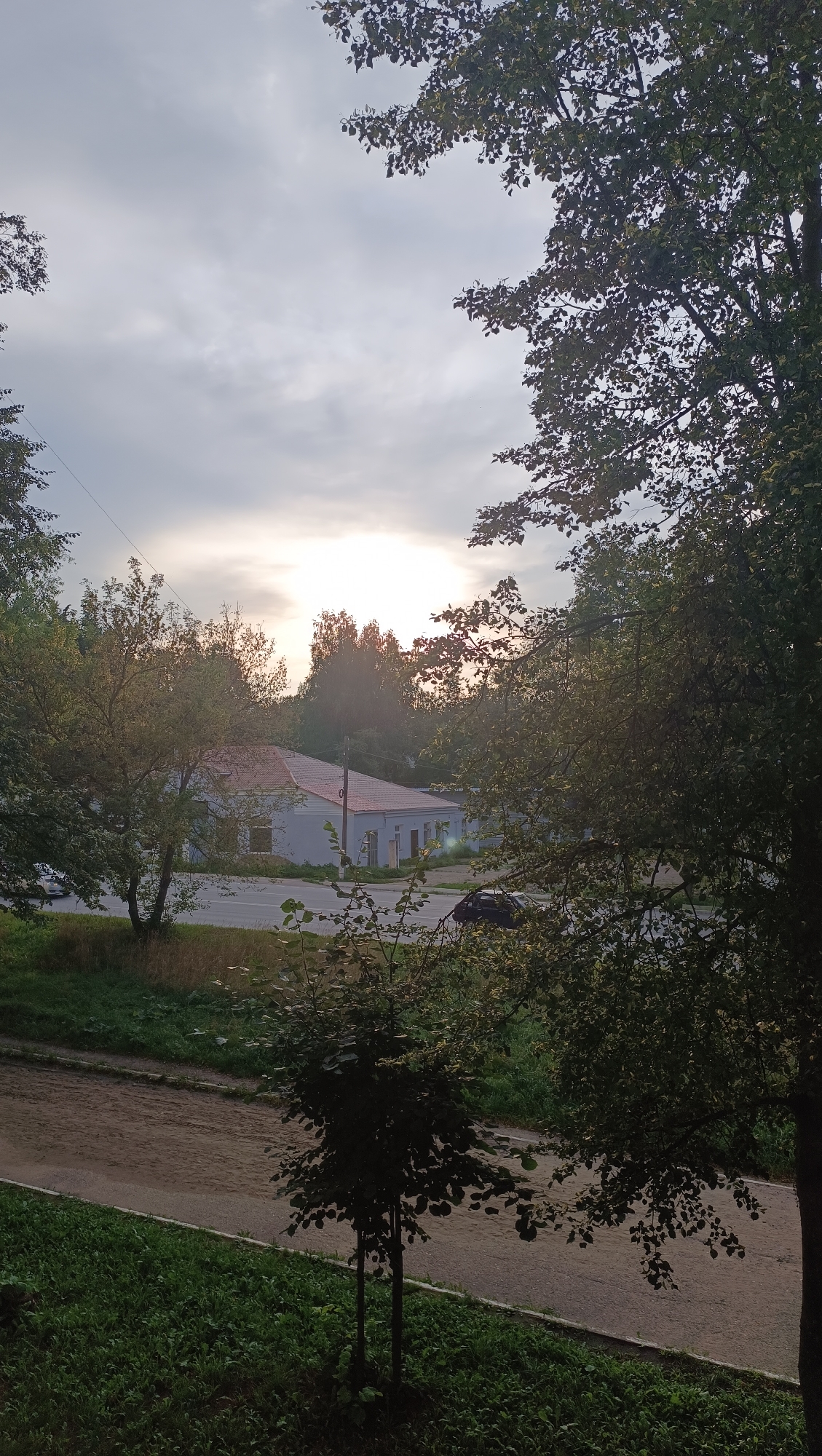 An overcast sky with the sun breaking through is visible above a low-rise building with a reddish-orange roof, partially obscured by trees. The building appears to be a single-story structure with windows and a white facade, situated along a road. On the road, a dark-colored car is parked or moving. The foreground features a dirt path and lush green grass, with a small, silhouetted tree in the middle distance. The scene captures a suburban or semi-rural environment, consistent with the outskirts of Chaikovsky, Russia, likely during the late afternoon or early evening given the lighting.