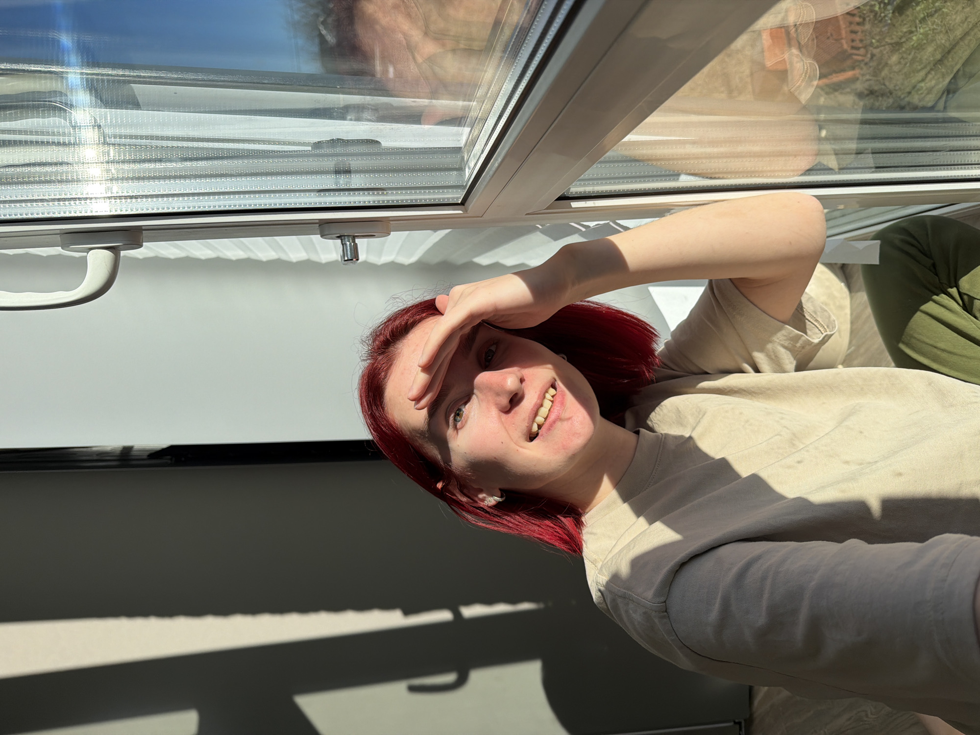 A young woman with short, bright red hair stands indoors in front of a window. She is wearing a beige t-shirt and green pants. She is smiling and her hand is raised to her forehead, shielding her eyes from the sunlight streaming through the window. The window has white frames and horizontal blinds. The reflection in the window is of an indistinct indoor scene. A shadow from the window frame is cast on the wall behind her. The location is Chaikovsky, Russia.