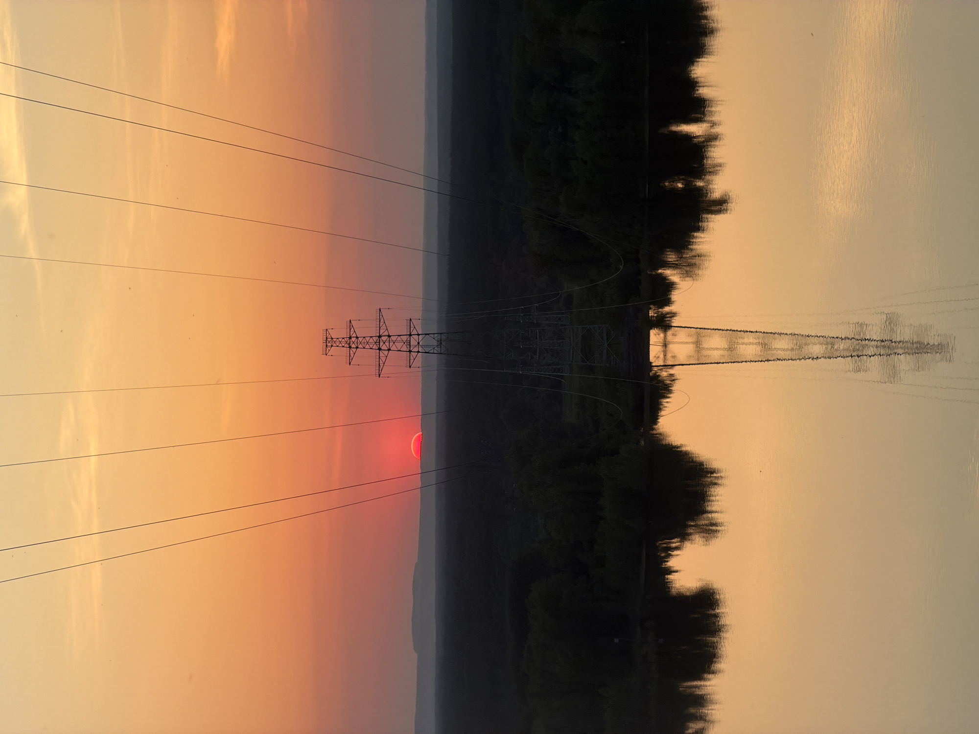 The image presents an inverted landscape dominated by a sunset over a body of water, likely the Volga river. A high-voltage power line structure stands prominently in the midground, reflected in the calm water below. The setting sun appears as a partially obscured red disc behind the power line. Overhead power lines extend across the frame, creating a grid-like pattern against the sky. The background shows a dark silhouette of vegetation along the water's edge. The scene exudes a sense of tranquility despite the industrial presence, under an orange and yellow hue of the sunset, likely in the vicinity of Noviy, Russia.