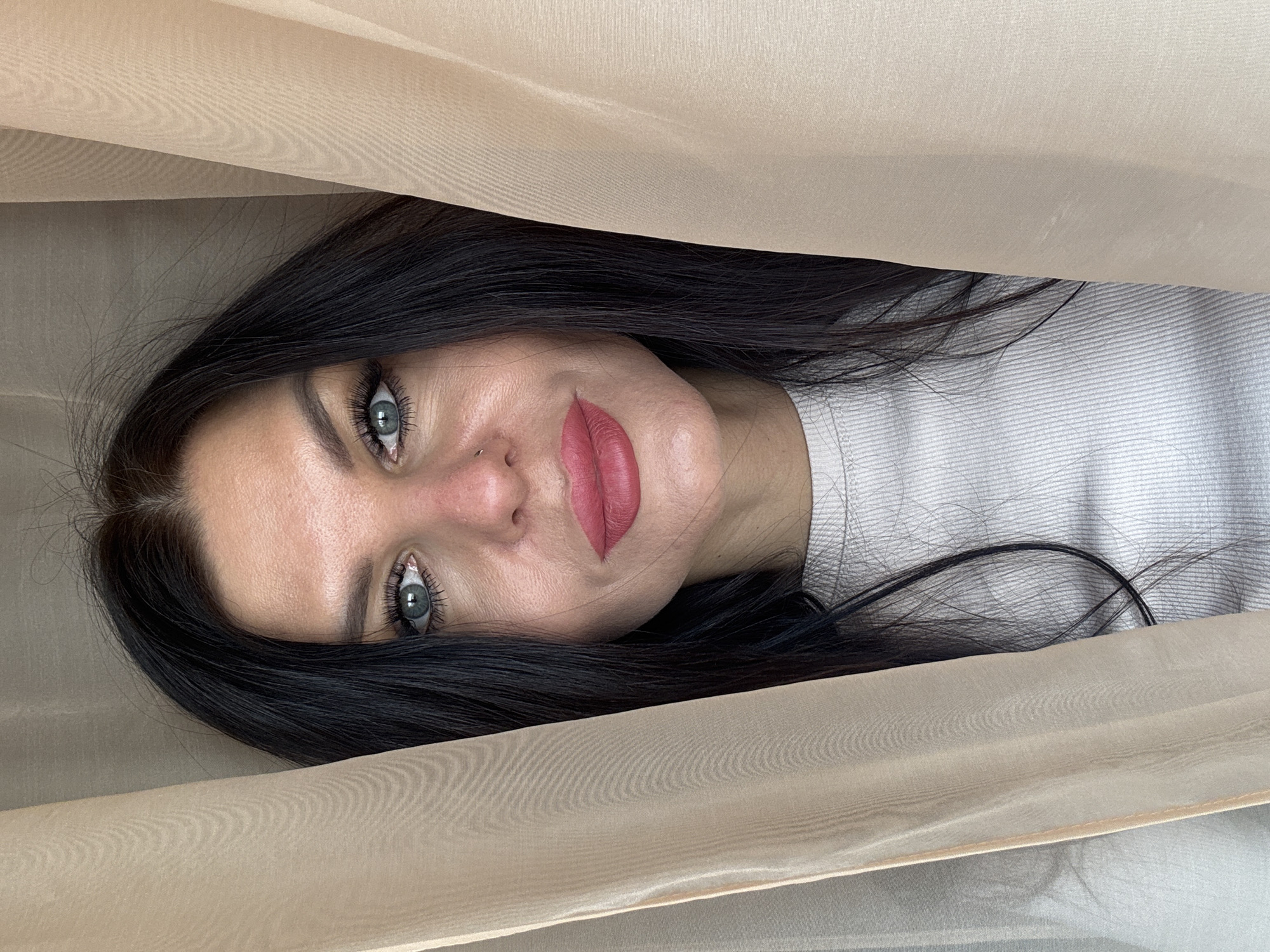 A woman with dark hair and light eyes is looking at the camera, framed by a translucent beige fabric. She wears a light-colored top. Her lips are painted red. The background is out of focus. No details indicate the location is Perm, Russia.