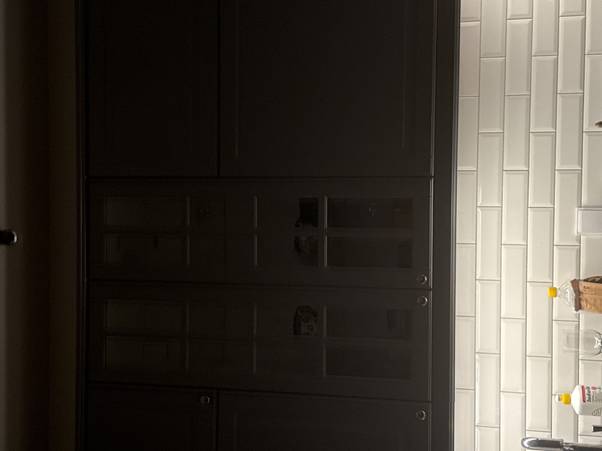 This is a medium shot, eye-level photograph of a kitchen counter area. The counter is covered in white subway tiles, and a faucet is visible on the left side. In front of the tiles, there are two bottles and a glass. One bottle is white with red and black lettering, and the other is clear with a yellow cap and a brown label. Above the counter are dark gray kitchen cabinets. The cabinets have glass doors on the left side, revealing some blurry contents within. The overall lighting in the image is dim. The location is listed as Unknown, Unknown.