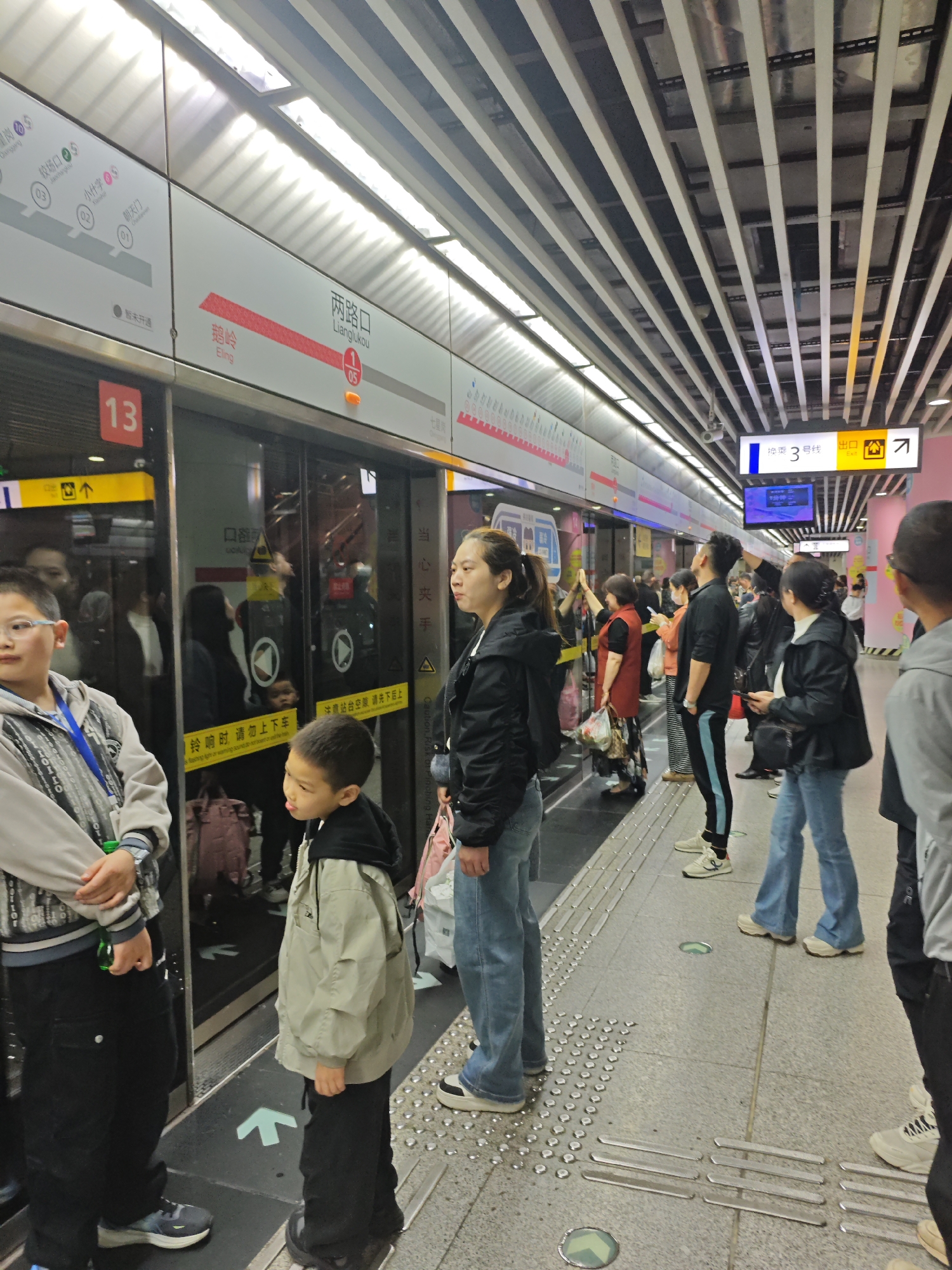 The image shows a subway platform in Yuzhong District, China. Several people are waiting near the subway doors. The doors are marked with the number "13" and have signage for "Lianglukou" station, indicating Line 1 and Line 05. There are directional arrows on the platform floor. Overhead signs indicate "Line 3" and an exit. The platform has tactile paving. The subway interior is visible through the doors, reflecting faces. The lighting is provided by long, linear fixtures.
