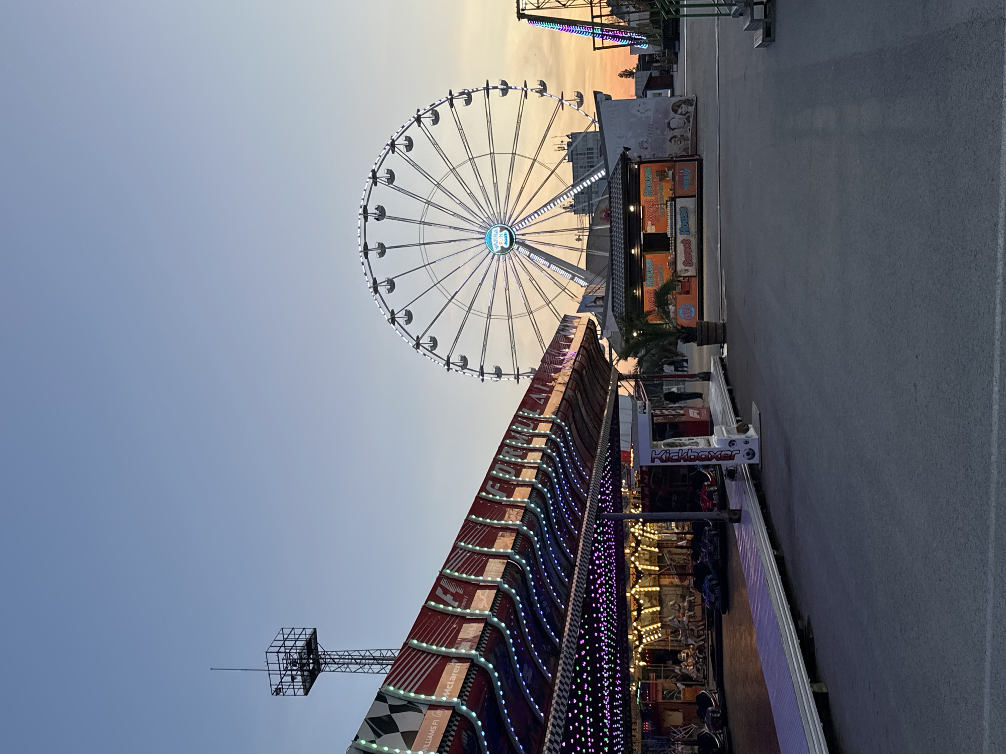 The image depicts an amusement park scene likely in Varna, Bulgaria, evidenced by the visible Ferris wheel known as "The Eye of Varna". The foreground showcases a carousel ride with multiple lights. A structure with red and white stripes along with smaller colored lights dominates a large portion of the image. The background presents a sky that transitions from light blue to orange-yellow hues, suggesting either sunrise or sunset. To the right of the Ferris wheel, there's a food stall or shop. The ground is paved, indicating a road or walkway within the amusement park. A small palm tree is near the stall. A structure with the signage "Kickboxer" is apparent near the right part of the carousel. There are people present in the lower portion of the carousel structure and near the stall. Overall, the scene captures a lively amusement park with lights and attractions.