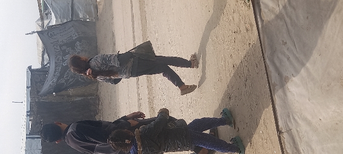 A group of people stand in an outdoor area. A young woman with long hair, wearing a gray patterned jacket and dark pants, walks past temporary dwellings. Another person walks ahead with a dark jacket and patterned pants, their back to the camera. Behind them, a male figure in a dark jacket looks on. The environment appears to be a temporary settlement, with makeshift shelters nearby.