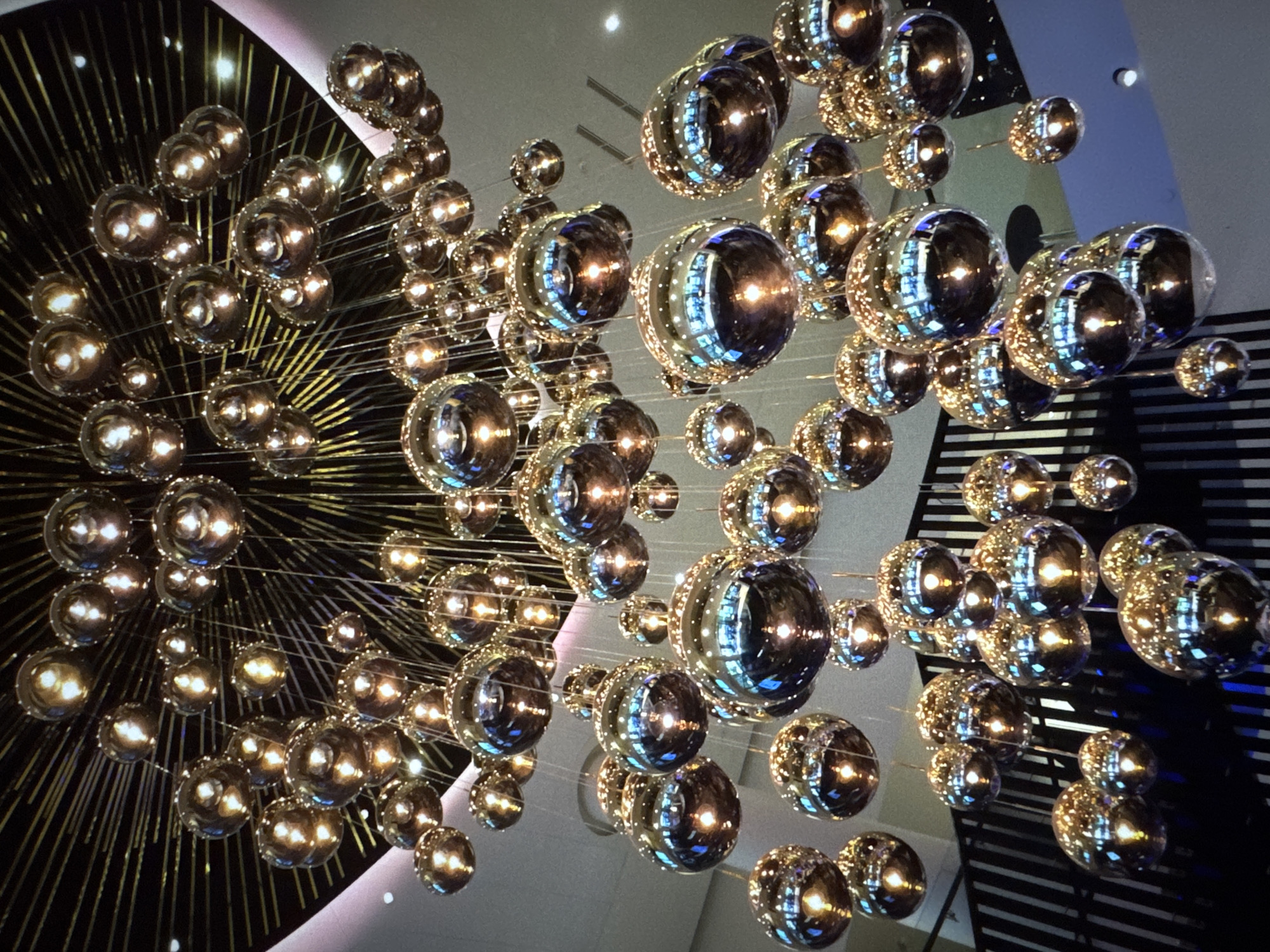 The image shows a large, modern chandelier made of numerous reflective spherical bulbs suspended from a ceiling. The bulbs vary in size and are arranged in a cascading, organic manner. Some bulbs are illuminated from within, casting a warm glow. The chandelier is suspended above a staircase with dark railings and treads, suggesting it is in a public space or a well-decorated interior. The background includes elements of the ceiling and walls, with soft lighting. There are no people visible in the image. The scene appears to be an interior architectural detail, likely in a hotel lobby, a shopping mall, or a similar establishment.