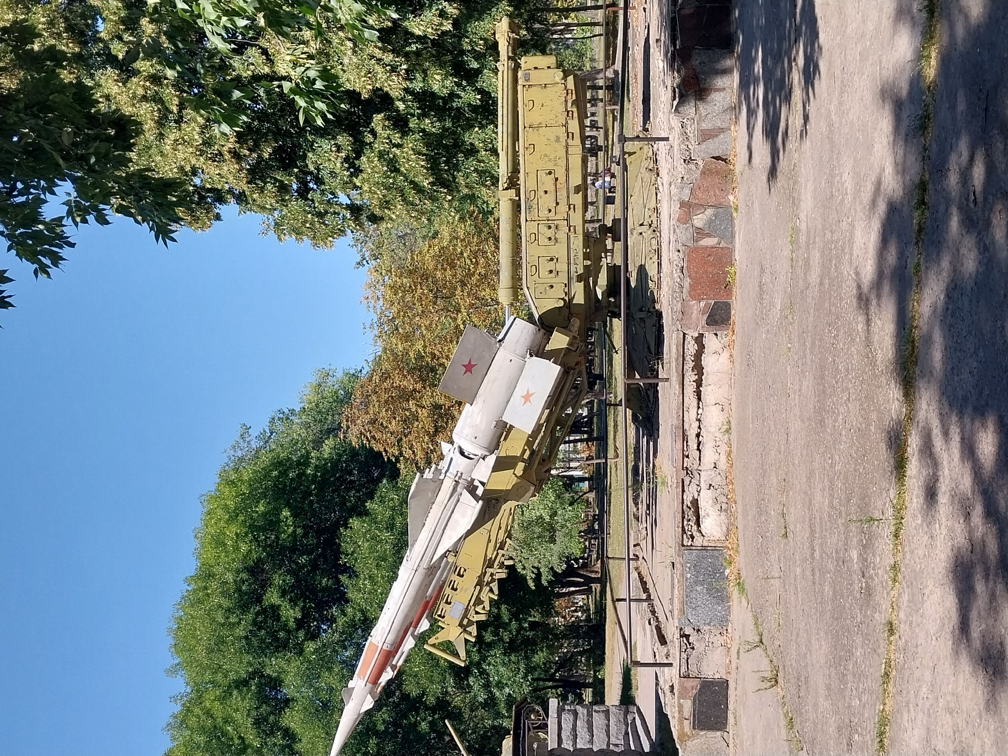 An outdoor exhibit, likely a museum or memorial, features a Soviet-era surface-to-air missile system on display. The missile is mounted on a wheeled launcher painted olive green, which itself rests on a concrete and stone foundation. The missile, primarily white with red and orange accents, is angled upwards, suggesting a ready-to-fire posture. Behind the missile system, lush green trees form a backdrop under a clear blue sky. The ground in front is paved with concrete, with patches of grass growing through cracks. In the distance, a few people can be seen near the exhibit, and additional trees and foliage suggest a park-like setting within the city of Dnipro, Ukraine.