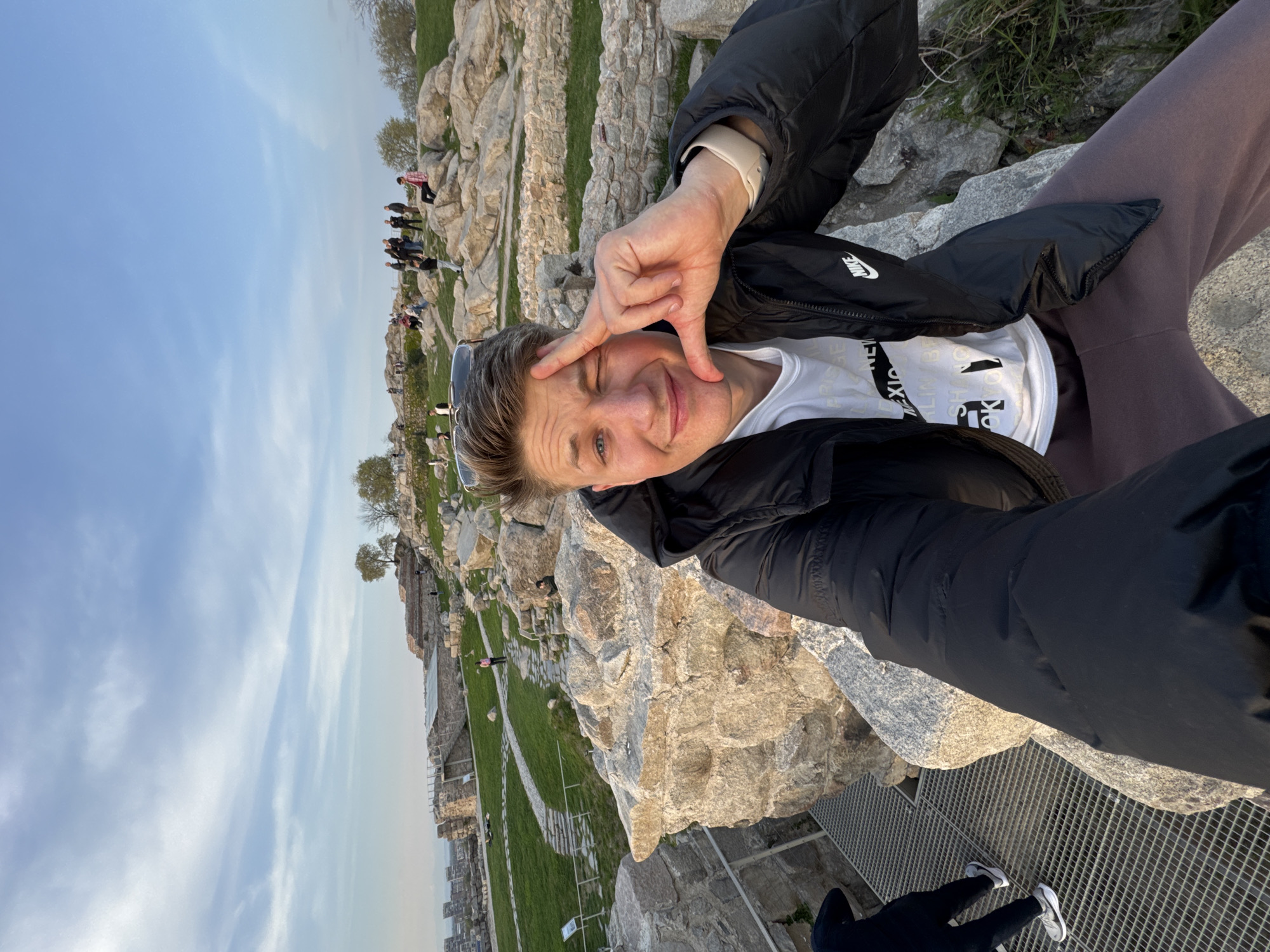 A man takes a selfie outdoors at Nebet Tepe in Plovdiv, Bulgaria. He is fair-skinned, with short blonde hair, and wearing a black jacket over a white t-shirt, and brown pants. He makes a hand gesture with his right hand, framing his face, and winks at the camera. Sunglasses rest on top of his head. He is standing on a metal grate. Behind him, a cliff of light-colored rocks overlooks a green landscape with ancient ruins and a city in the distance. Other people are visible in the background. The sky is blue with white clouds.