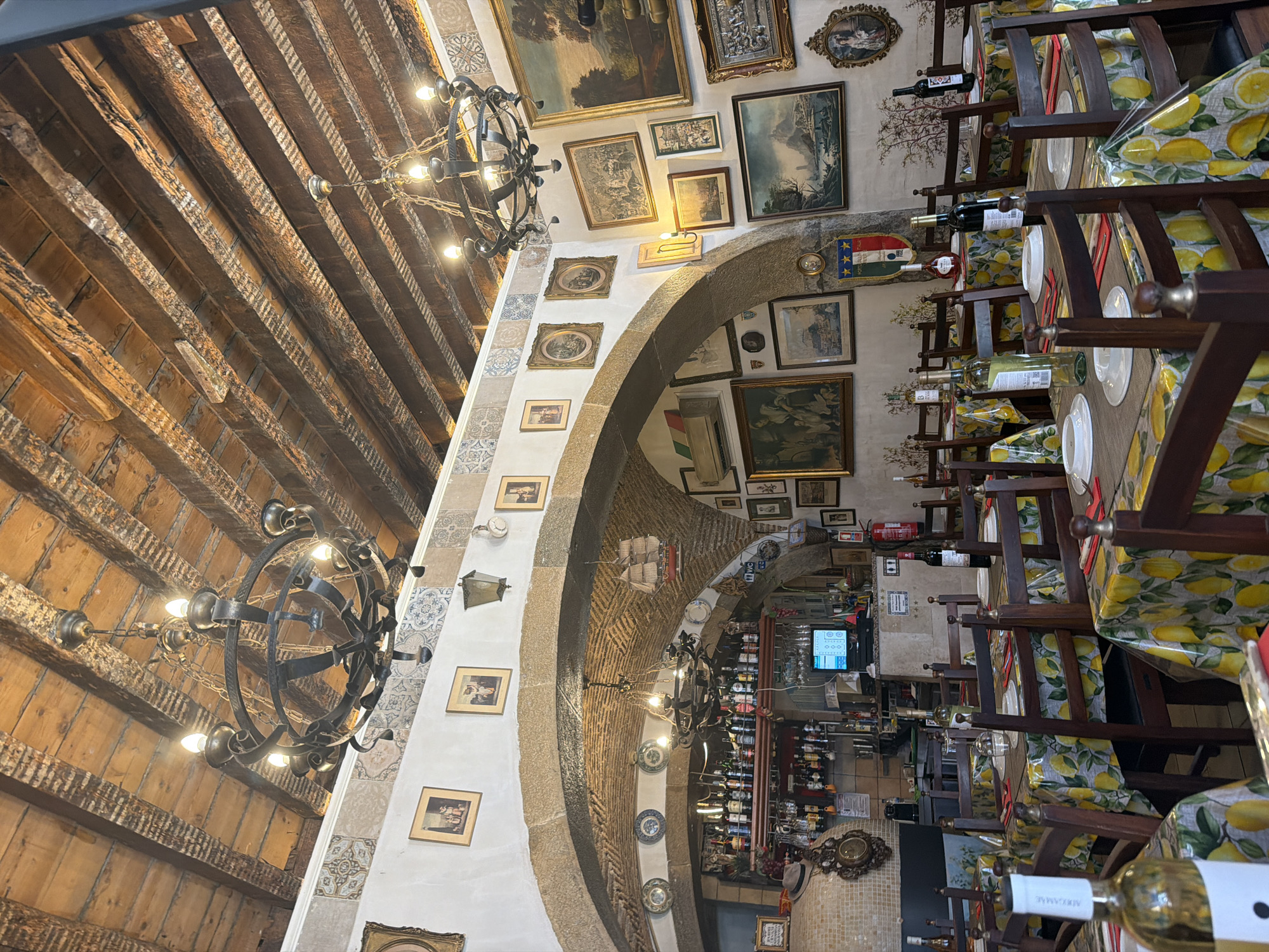 The image shows the interior of a restaurant with wooden beam ceilings and exposed brick arches. The tables are set with yellow, lemon-patterned tablecloths, wooden chairs, and white plates. There are multiple paintings and decorative plates on the walls. A bar area is visible in the background, stocked with bottles of alcohol. Two large, wrought-iron chandeliers hang from the ceiling. The restaurant appears to be empty of patrons. The presence of an Italian flag and a sign with "WC" suggests a connection to Italy, but the location is not specified as Lisbon, Portugal.