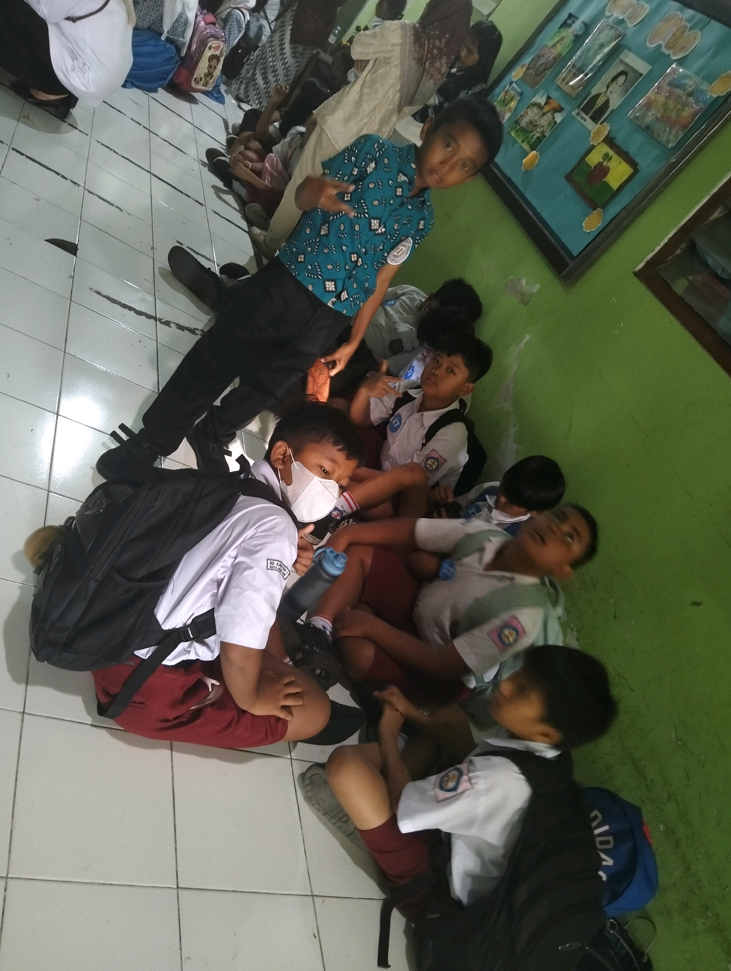 In a classroom setting in Magelang, Indonesia, several Indonesian school children, likely elementary or middle school age, are seated on a tiled floor next to a green wall. They are wearing school uniforms consisting of white shirts with school emblems and either maroon or dark colored shorts. Most of the children have backpacks, varying in color from black to blue. One child in the foreground is wearing a white face mask. The children are arranged in a cluster, some are looking at the camera, while others are looking away or interacting with each other. One child is holding a water bottle. On the green wall behind them, there is a display board with several framed pictures or drawings, possibly student artwork. The lighting suggests an indoor environment, possibly during daytime.