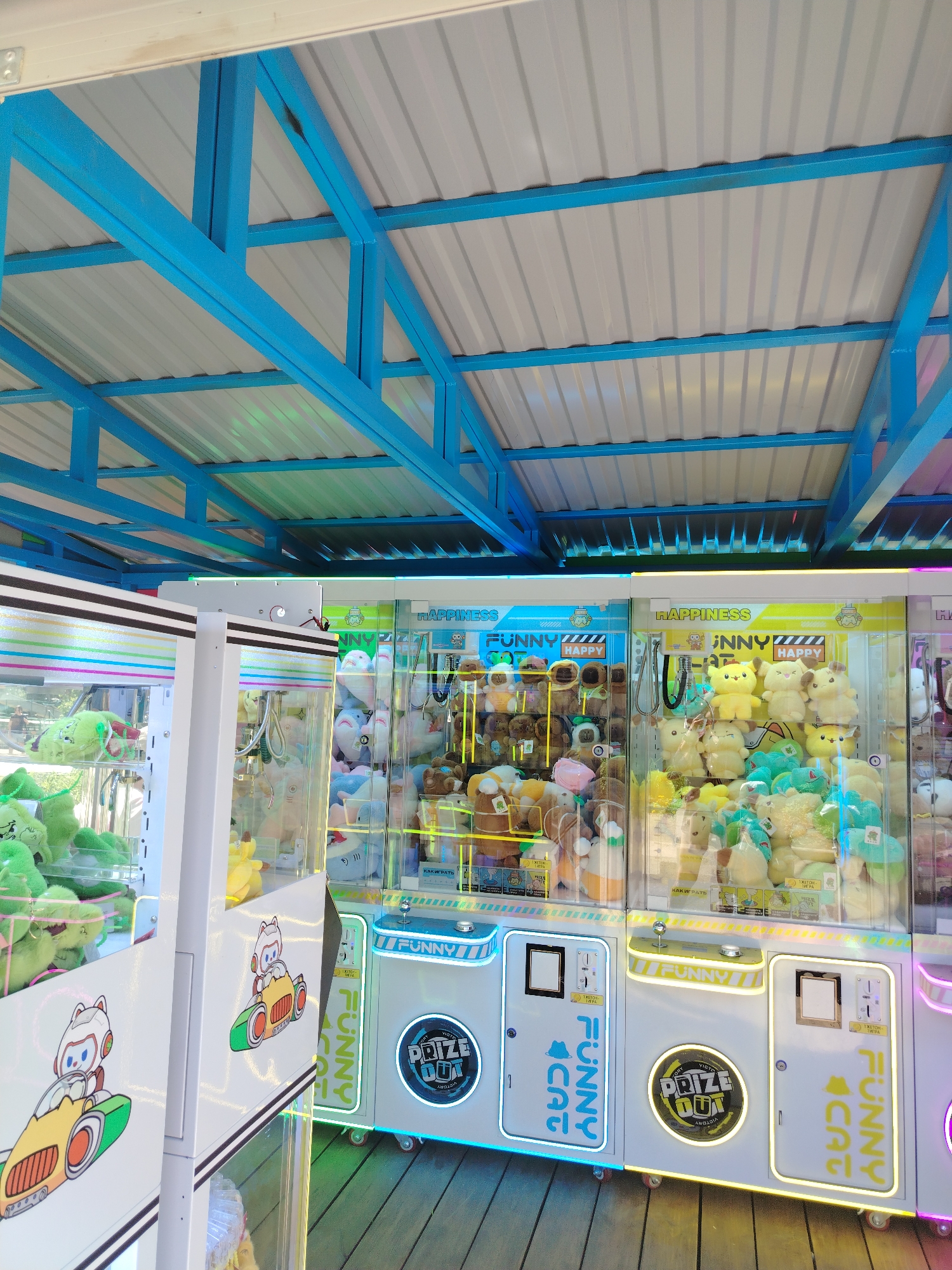 The image captures a row of claw crane arcade games. The machines have a white casing with neon-colored accents and clear glass displays filled with plush toys. The game to the left is filled with green plush animals. The center-left game is filled with brown stuffed animals. The center game is filled with yellow and green plush animals. There is some writing on the games in English, but also a Russian phrase "КАК ИГРАТЬ" meaning, "How to play". The machines sit on a wooden plank floor under a blue metal structure of beams and a white metal roof. The location is likely in the city of Chaikovsky, Russia, based on the presence of the Russian text.
