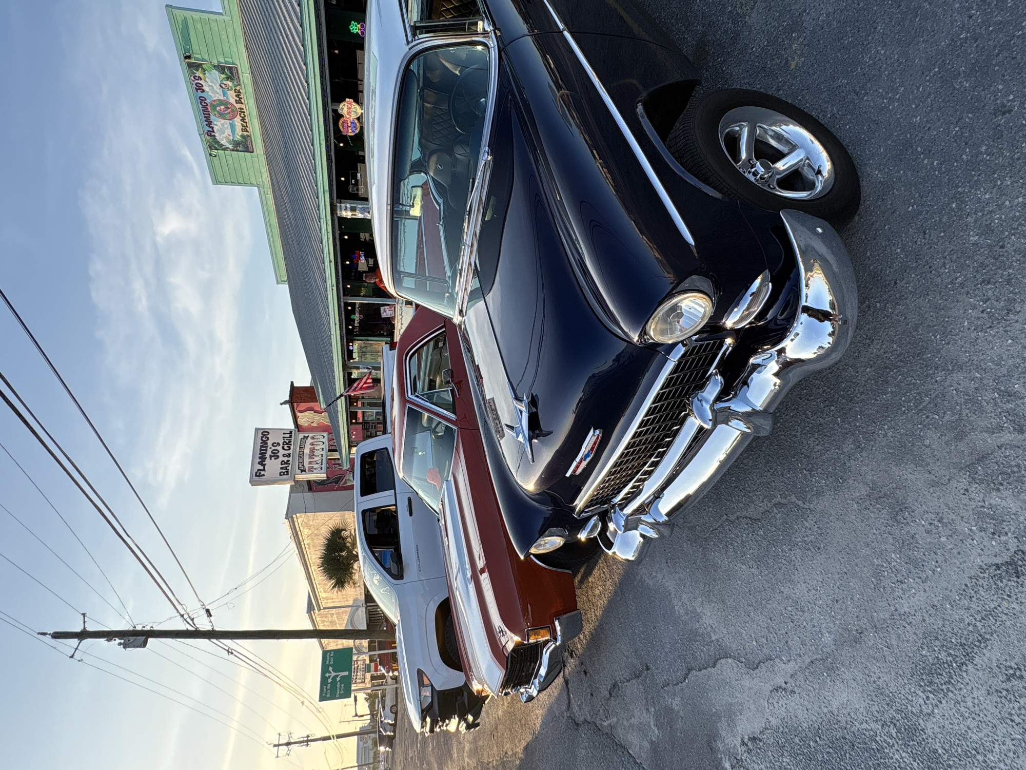 The image displays a vibrant street scene in Panama City Beach, United States of America, captured during what appears to be the late afternoon or early evening, indicated by the warm, directional light and extended shadows. The sky is clear with only a few wispy clouds.
In the foreground, a highly polished dark blue (or possibly black) classic car, featuring prominent chrome bumpers, grille, and trim, is parked on the asphalt street. Its custom chrome wheels catch the sunlight. Behind it, a second classic car, painted in a deep red or maroon hue, is visible, followed by a white Chevrolet Colorado pickup truck. These vehicles suggest a car gathering, show, or simply regular parking in a popular area.
To the left of the parked cars stands a building with light green siding, housing "FLAMINGO JO'S BEACH BAR," as clearly advertised on a large sign depicting a beach scene. Further along the building, another sign identifies it as "FLAMINGO JO'S BAR & GRILL AMERICAN FOOD." The establishment appears to be a casual eatery or drinking spot.
The upper portion of the scene is crisscrossed by multiple utility poles and power lines against the bright sky. A green road sign, partially obscured but legible, provides directions for local roads including "Front Beach Road," "Middle Beach Road," "South Thomas Dr," and "Hutchinson Blvd," unequivocally confirming the Panama City Beach setting. A small palm tree adds to the coastal atmosphere. The overall impression is one of leisure and classic American culture in a popular beach destination.