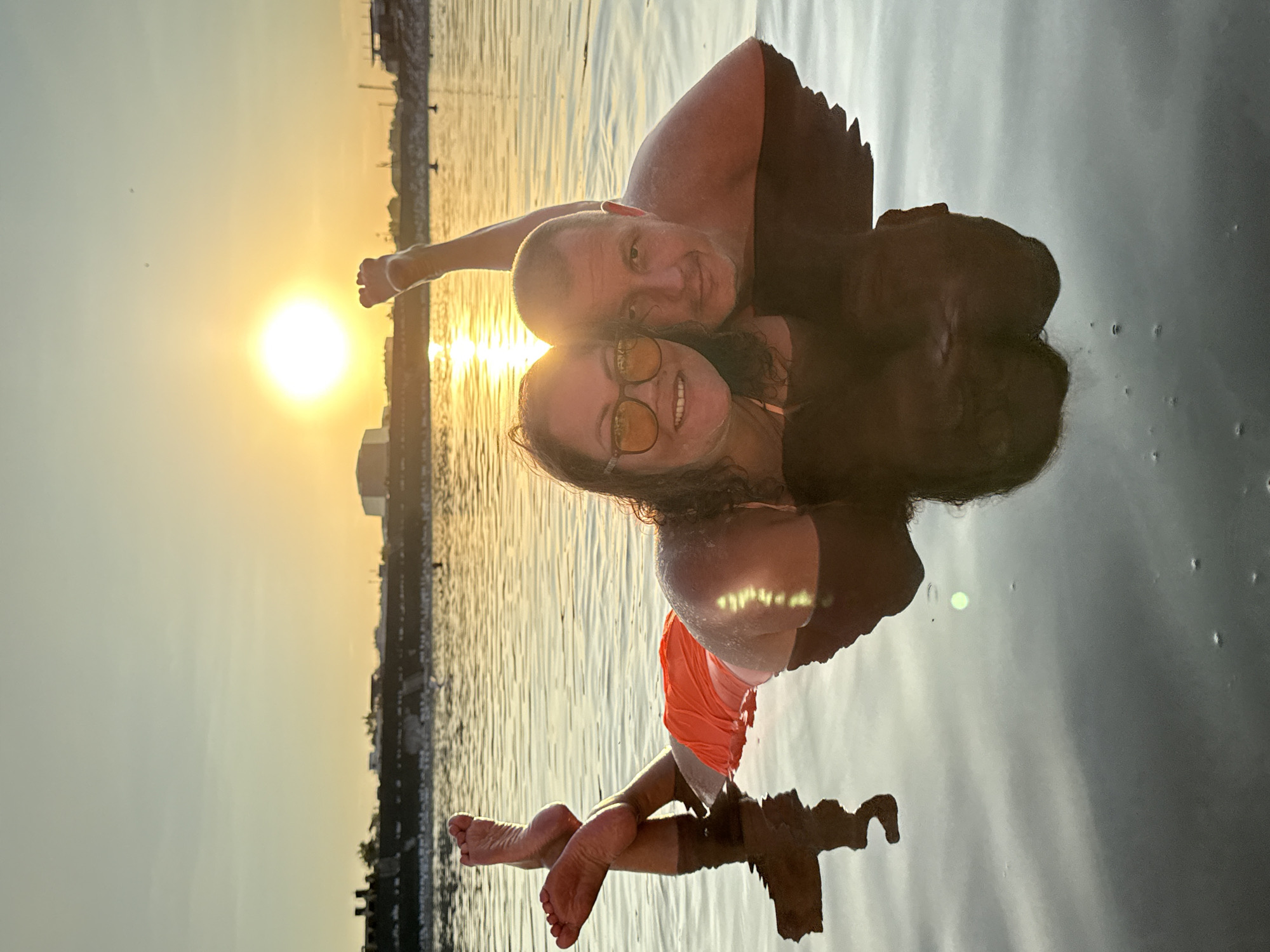 The image shows a couple lying in shallow water at sunset. The man and woman are facing the camera, with the woman in the foreground wearing sunglasses and an orange swimsuit. The man is behind her, looking directly at the camera. Both are partially submerged in the water, with their reflections visible below them. The sun is setting on the horizon, casting a warm glow on the scene and creating a strong lens flare. In the background, there is a city skyline with buildings and structures along the shore. The water appears calm and reflects the sky and the couple. The overall atmosphere is serene and romantic. This scene is likely captured in the city of Sol-Iletsk, Russia, known for its salt lakes and recreational areas.