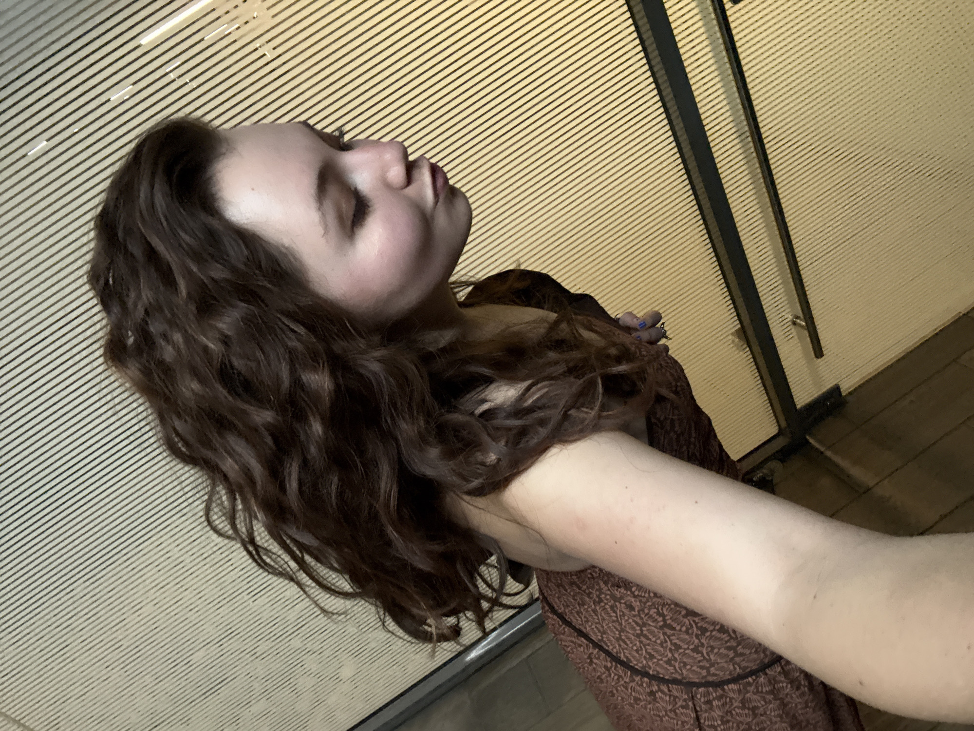 A young, fair-skinned woman with long, wavy brown hair is posing in front of what appears to be the entrance to a subway station. She is wearing a patterned brown dress. The background is a cream-colored wall with horizontal lines, and a dark metal door frame is visible on the right side. The location is possibly in Mexico City, Mexico, based on the architectural style of the subway station entrance.