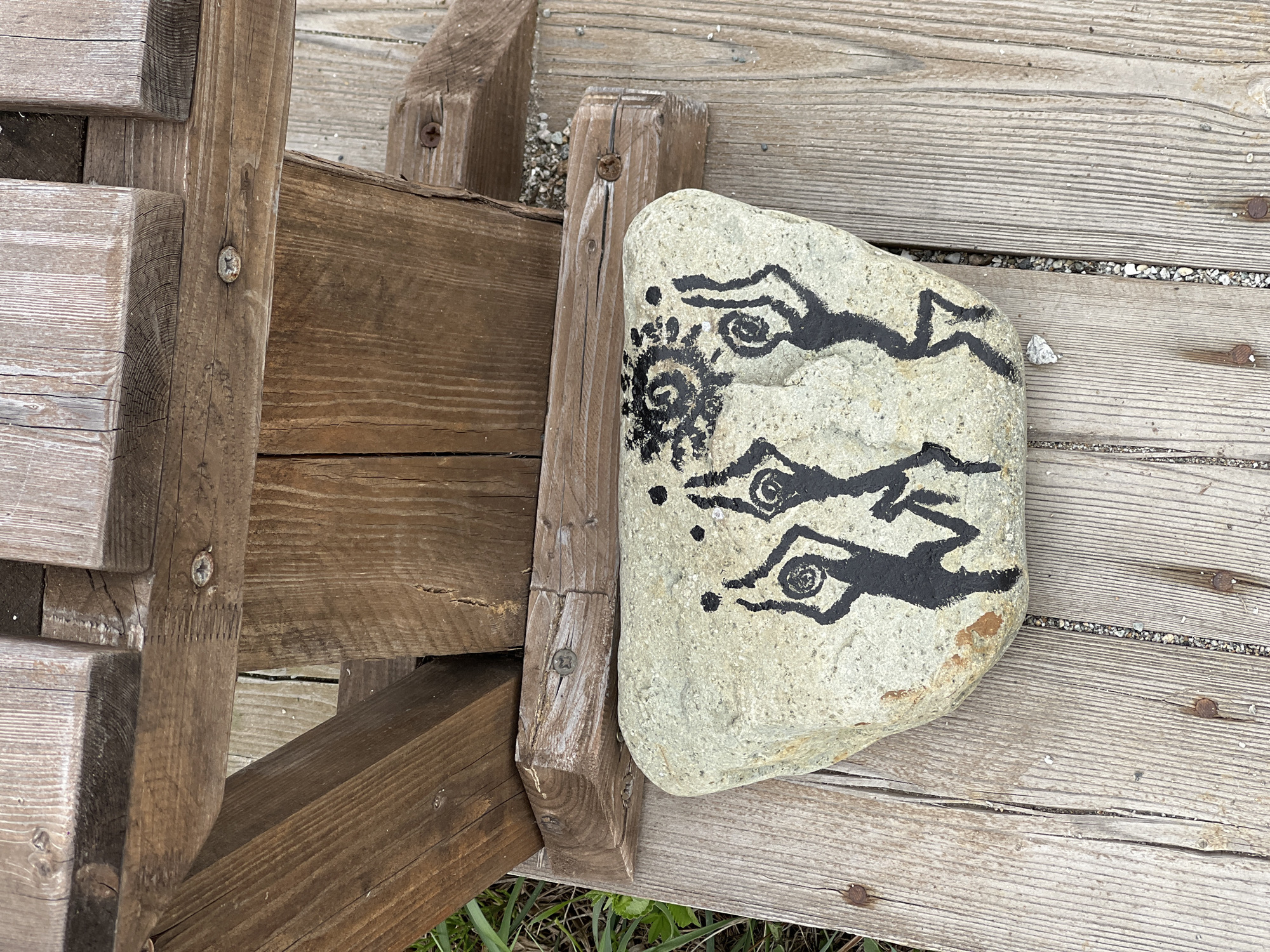 The image shows a light-colored, irregularly shaped rock with black drawings on it. The drawings include what appear to be stylized figures with large eyes and a circular design resembling a sun or spiral. The rock is sitting on a wooden surface with parallel planks, likely a deck or walkway. To the left of the rock is a section of wooden structure, possibly a fence or steps, constructed from thicker, darker wood beams. The environment suggests an outdoor setting, with some sparse grass visible in the lower portion of the image. There is no explicit information to definitively place the image in Zarubino, Russia; this detail is inferred from the instructions.