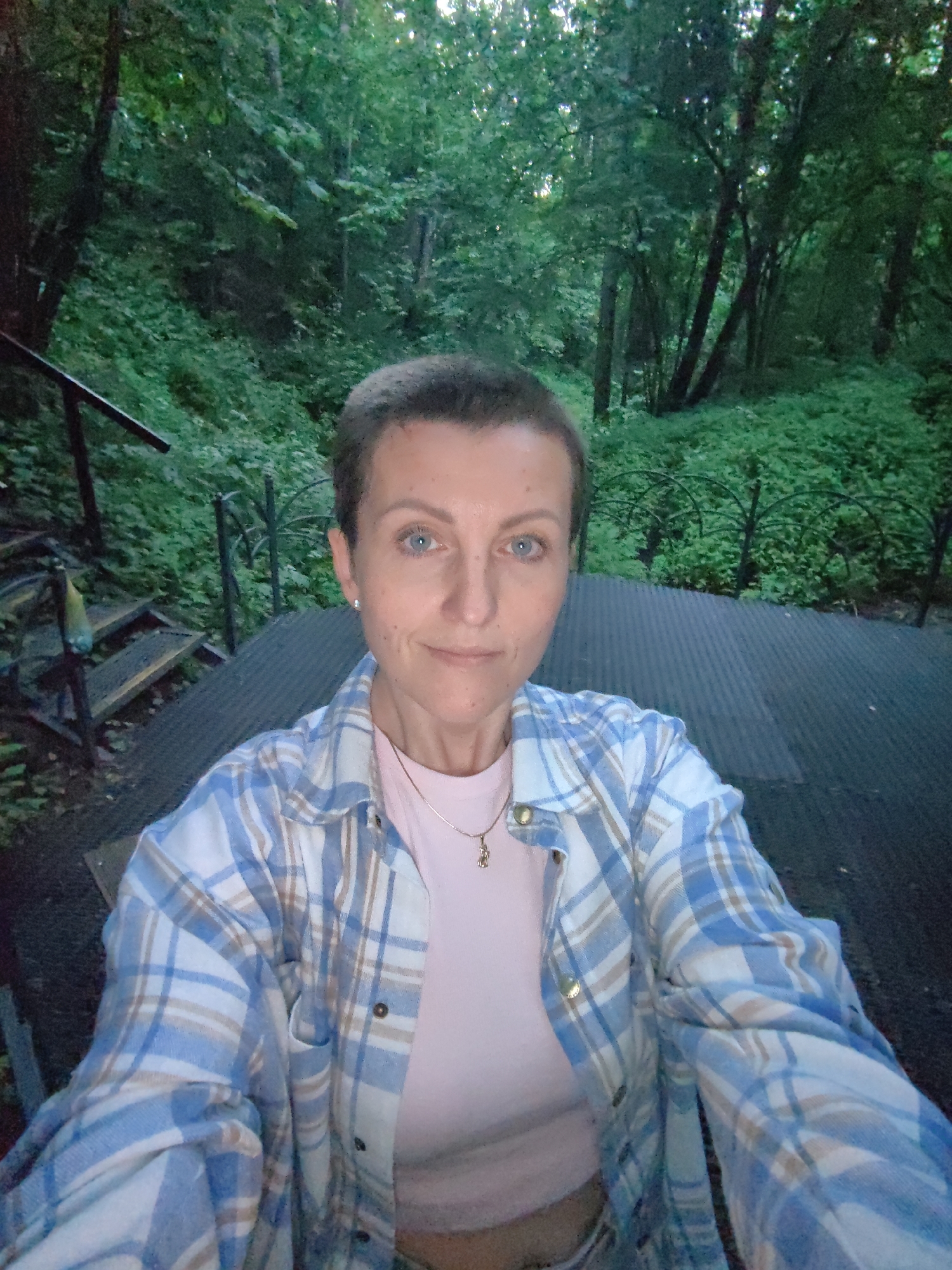 A woman with short brown hair and blue eyes is taking a selfie outdoors. She is wearing a pink t-shirt and a blue and white plaid shirt. She is standing on a dark, ribbed surface, possibly a bridge or walkway, with stairs and railings to her left. The background is a dense green forest with trees and foliage. The scene appears to be in a park or natural area within the Дзямідавіцкі сельскі Савет region of Belarus.