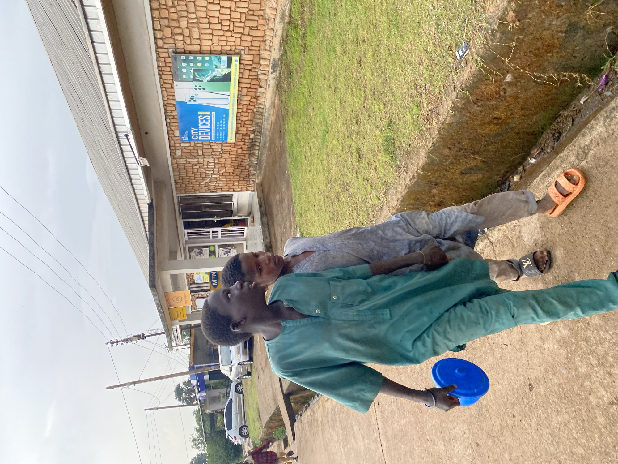 Two adolescent males are positioned on a sidewalk in Bukuru, Nigeria. One male is wearing a teal, long-sleeved button-down shirt, long pants, and holding a blue plastic container with a lid. He is looking upwards. The other male, wearing a light gray outfit, is lying on his back, and looking at the camera. One foot is bare, and the other has a black sandal with a gold symbol. The sidewalk is next to a patch of green grass. A building with a sign for "City Devices" is in the background. Cars are parked along the road. Overhead power lines are visible.