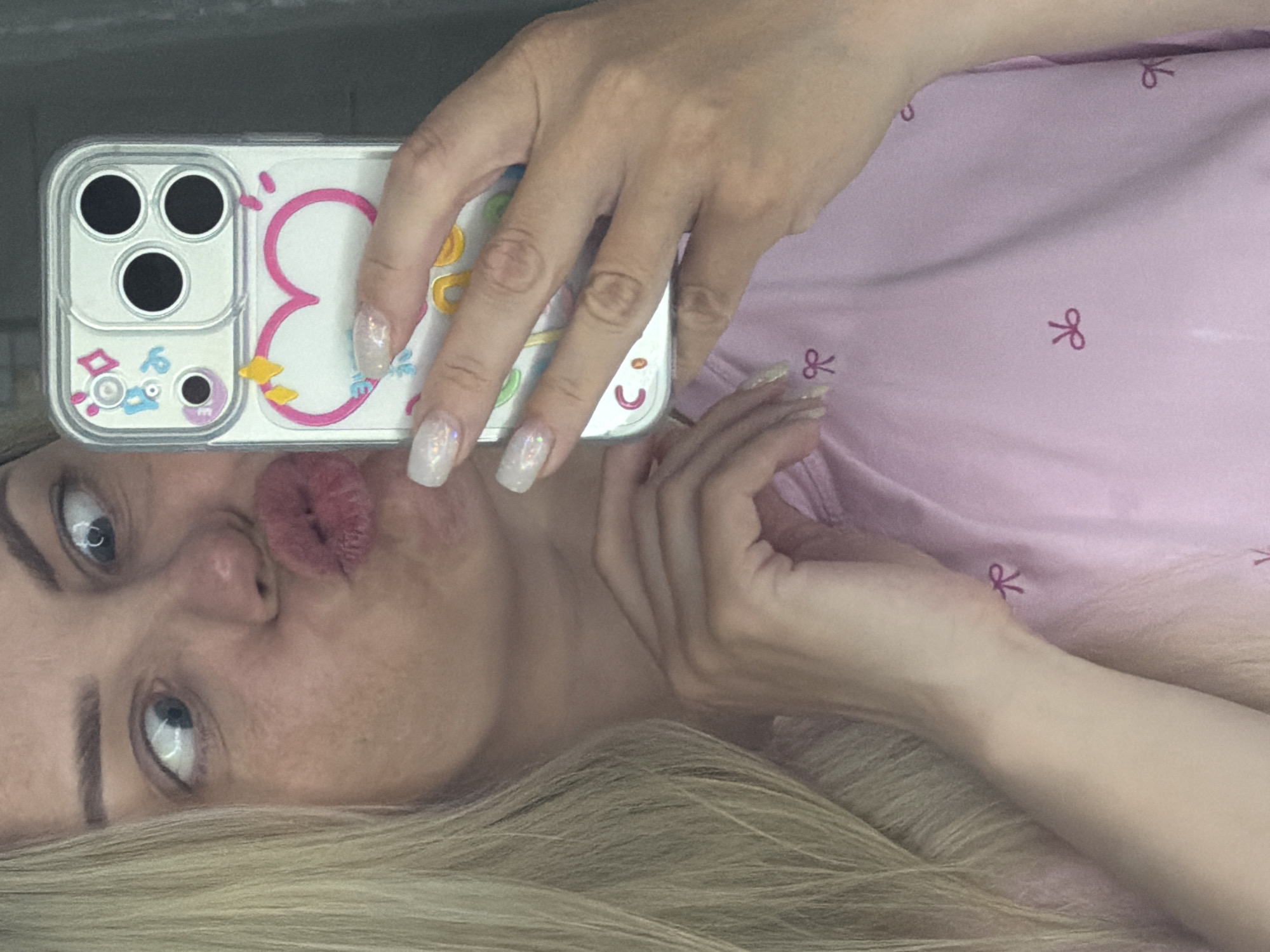 A person is holding a smartphone with a clear phone case with cartoon decorations. The person is making a kissing face towards the camera. They are wearing a light pink shirt with bow decorations. They have blonde hair and light-colored eyes. It is not possible to determine the location of the scene as Votkinsk, Russia from the image.