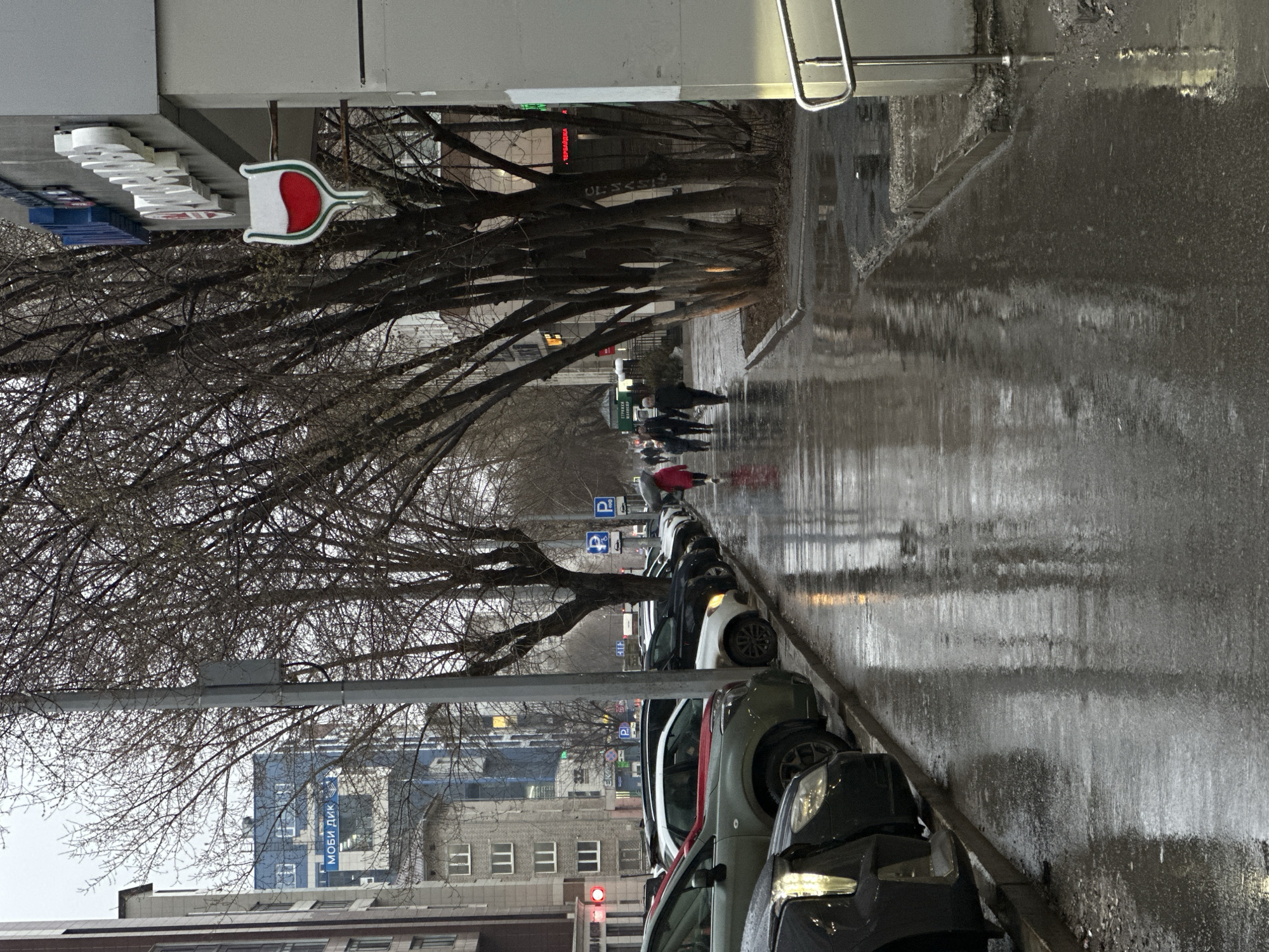 The image depicts a rainy street scene in Perm, Russia. The sidewalk is wet and reflective, indicating recent or ongoing rain. A row of parked cars lines the right side of the street, partially obscured by raindrops on the lens. Pedestrians with umbrellas walk along the sidewalk in the distance. Bare trees branch across the upper portion of the image, with some buildings visible in the background. A sign with a red and white symbol, likely for a business, is visible on the upper left. A building with a blue sign is discernible in the distance. The overall atmosphere is overcast and grey.