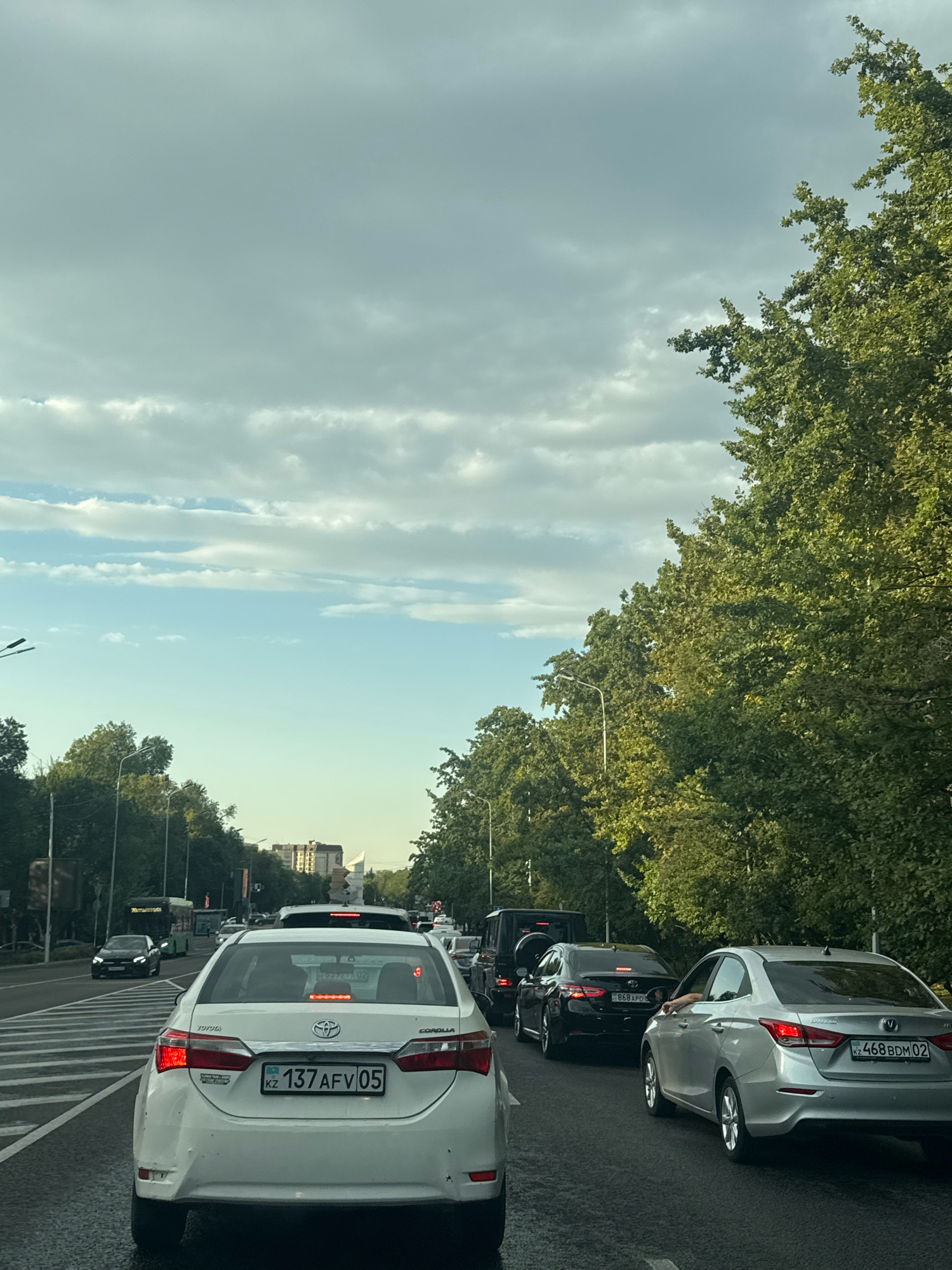 The image captures a traffic jam on a city street in Almaty, Kazakhstan. The perspective is from the driver's seat of a white Toyota Corolla, which is in the foreground, facing forward. The car has a license plate that reads "KZ 137 AFV 05". A silver Changan sedan with license plate "KZ 468 BDM 02" is to the right of the Corolla, and a black sedan with license plate "868 APD" is in front of it, followed by a black Mercedes-Benz G-Class. Multiple lanes of traffic are visible, with cars lined up. Lush green trees line the right side of the street, with more trees and buildings visible in the distance. The sky is overcast with clouds, and the light suggests it is either early morning or late afternoon. The road appears wet, possibly indicating recent rain.