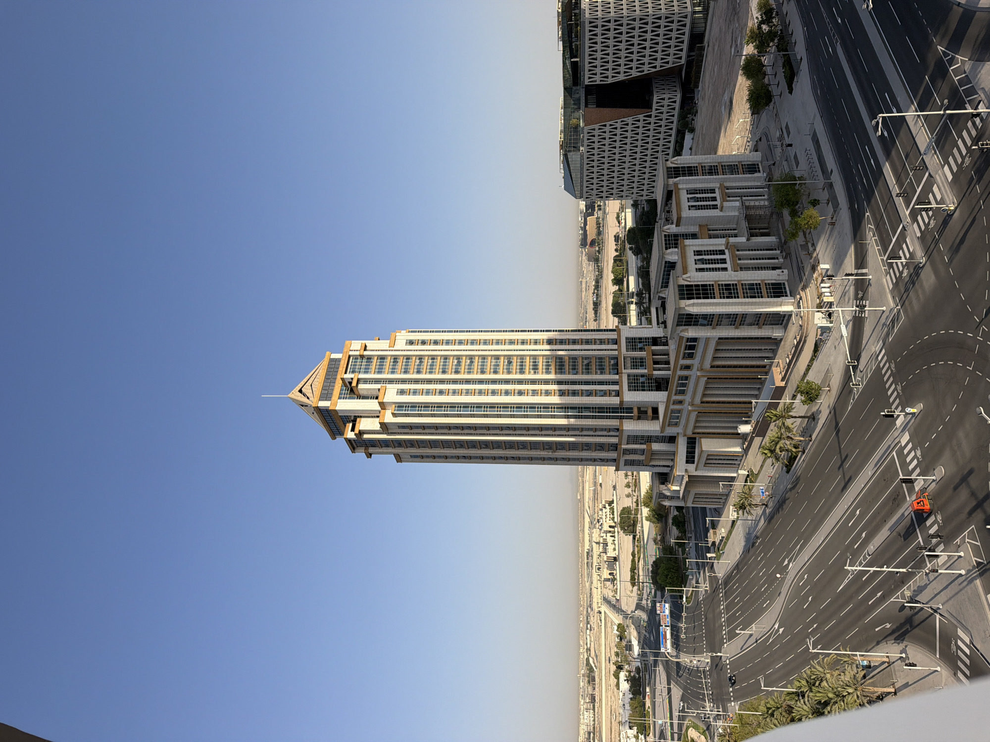 An aerial, eye-level, wide shot captures a modern, beige and glass skyscraper dominating the center of the frame. The skyscraper features a tiered design with a pointed, copper-colored roof. To the right of the skyscraper, a contemporary building with a white, geometric facade stands. The foreground showcases a multi-lane intersection with road markings and traffic lights, bordered by palm trees and sidewalks. In the background, a flat, arid landscape stretches towards a hazy horizon, dotted with low-rise structures and roads, indicative of an urban or developing suburban area. The sky is clear and blue. The scene is devoid of people and any overt action.