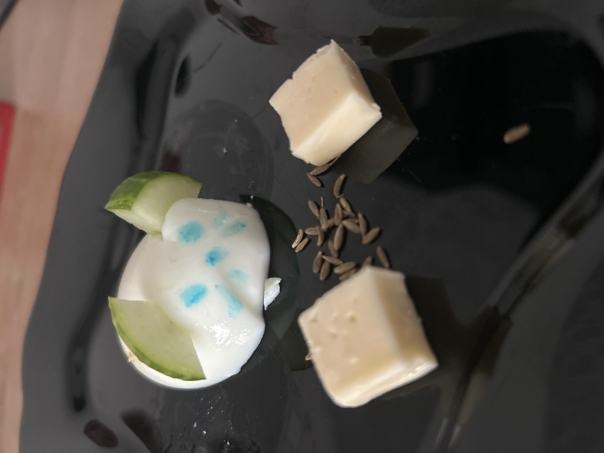 A black plate holds a small food arrangement. There are two cubes of a white, firm cheese, possibly Parmesan. Between them are about 10 dark brown seeds, likely fennel or cumin. Near the bottom left corner is a halved hard-boiled egg topped with a smear of white sauce and two cucumber slices. The sauce has several small blue spots. The plate is glossy and reflects light. The setting appears to be indoors with a surface that is reflecting a hint of red. The image doesn't contain information regarding the city of Chaikovsky, Russia.