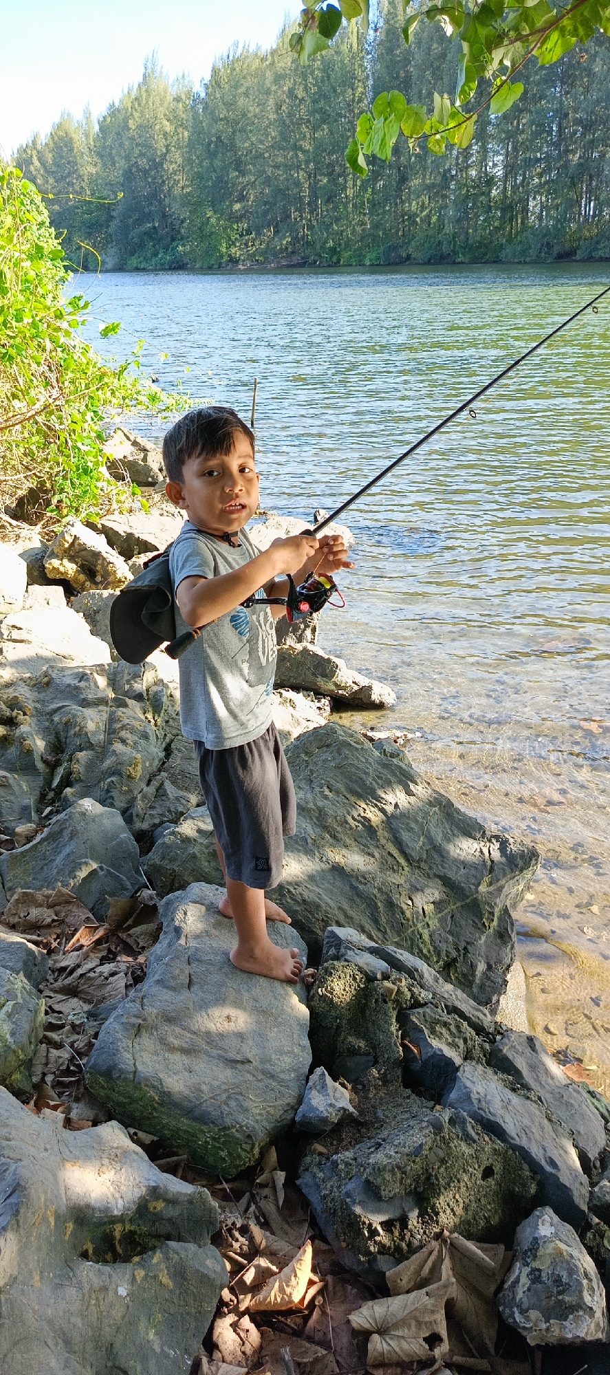 A young boy is fishing on the rocky shore of a body of water, likely a river or lake, in Meunasahmesjid, Indonesia. He is holding a fishing rod and reel, with the line extending into the water. The boy is wearing a grey t-shirt and dark shorts. He is standing on a large rock, with more rocks and dry leaves surrounding him. The background features a line of trees and the water, which appears calm with sunlight reflecting on its surface. The overall scene suggests a peaceful outdoor activity.