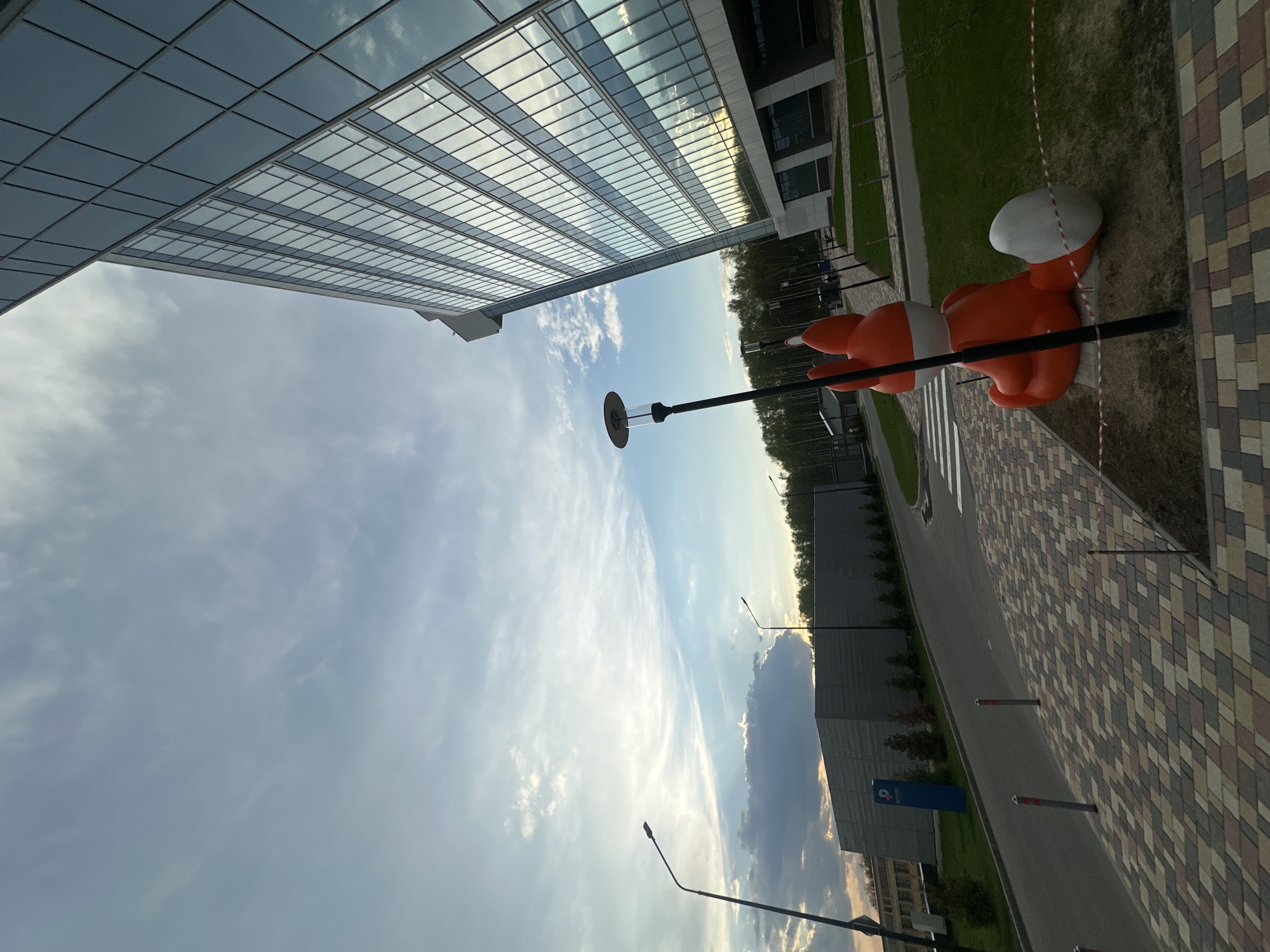 A building with a glass facade dominates the view of the sky. An orange fox sculpture is visible on a roadside in Moscow, Russia. The road is lined with a patterned sidewalk. Streetlights are seen along the road. A striped crosswalk marks the pedestrian path.